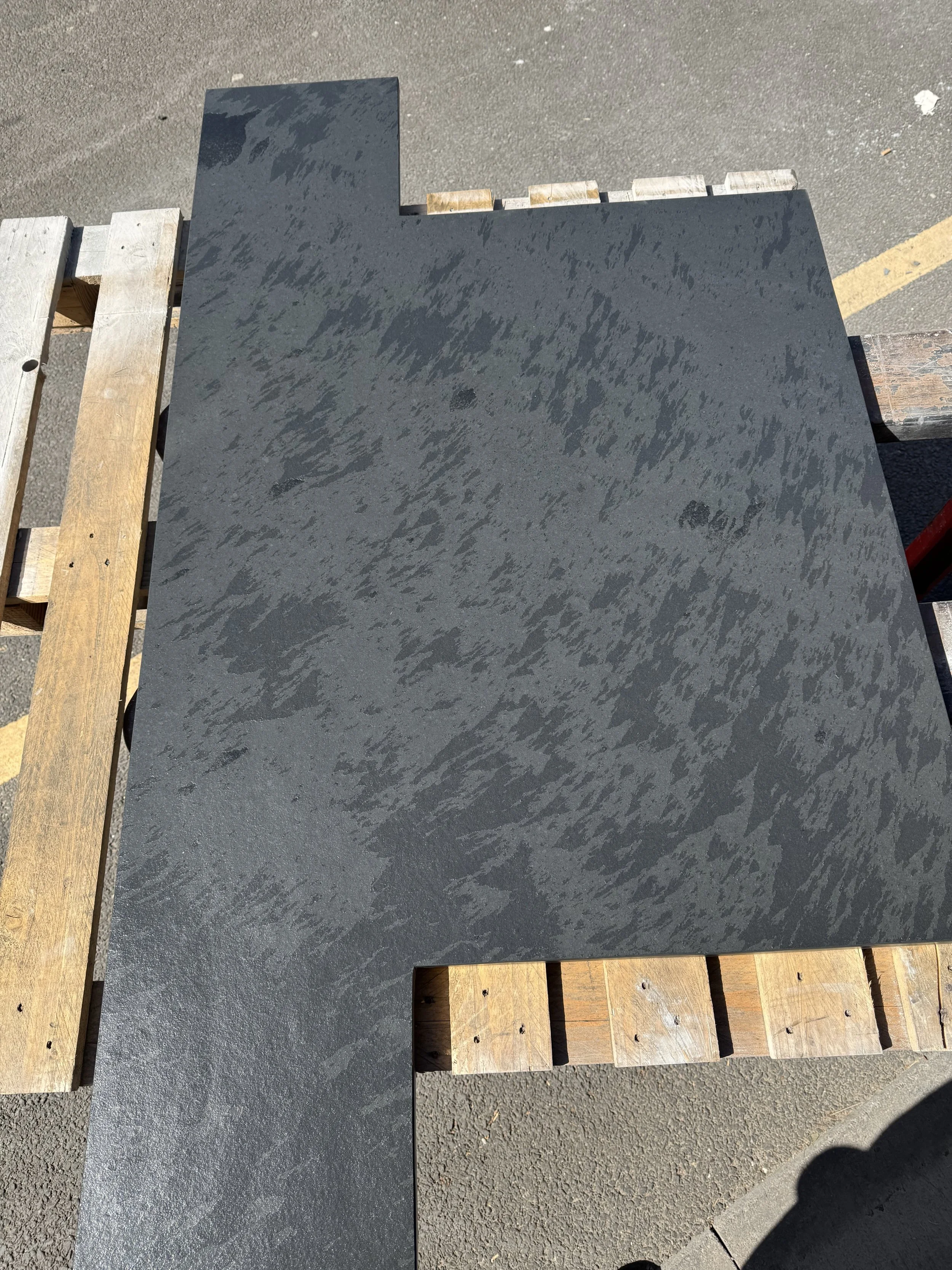 Black textured tile with an irregular L-shape design, placed on a wooden pallet outdoors.