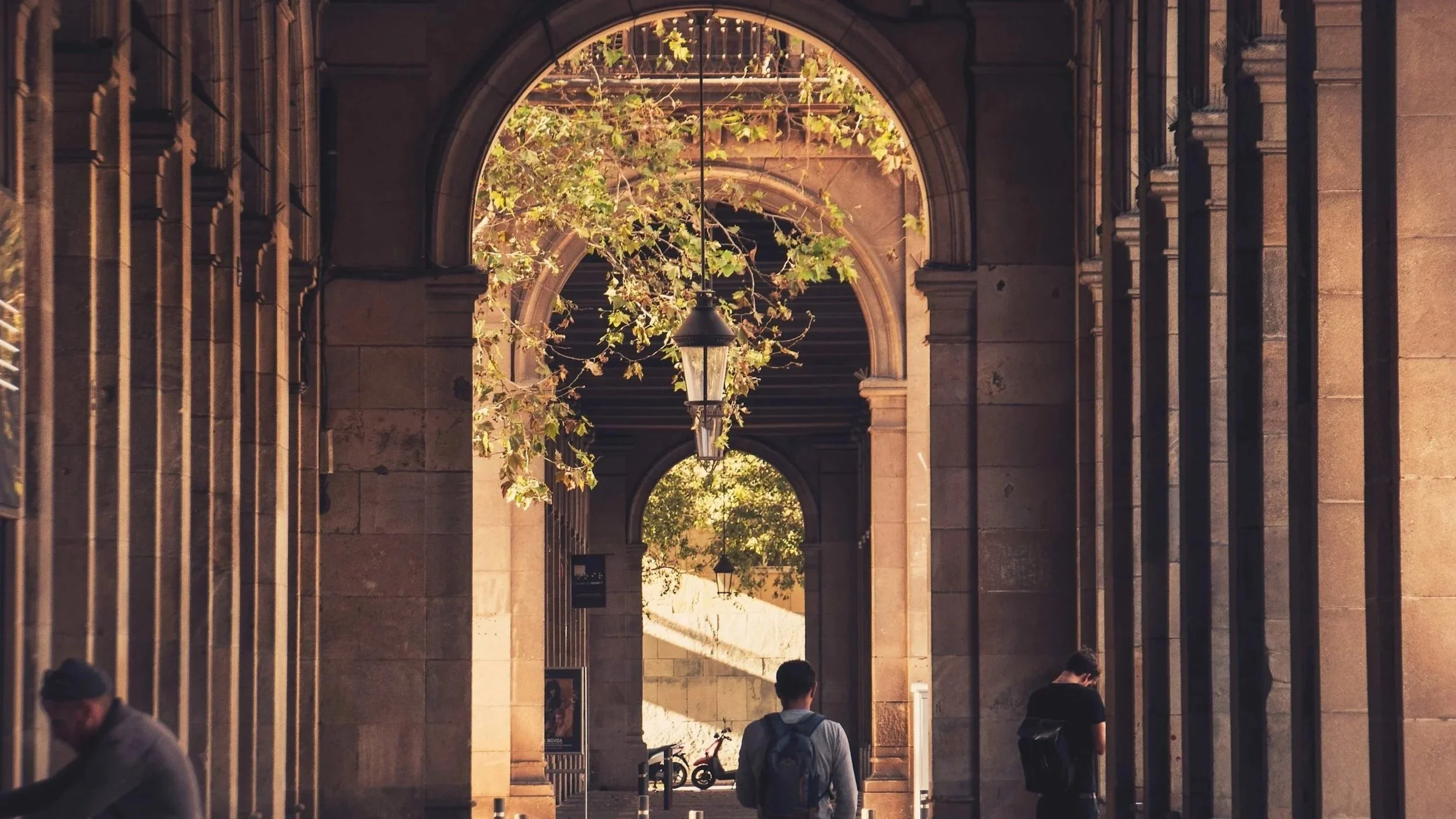 A colonnade in Barcelona with sunlight streaming in from the left side and people walking