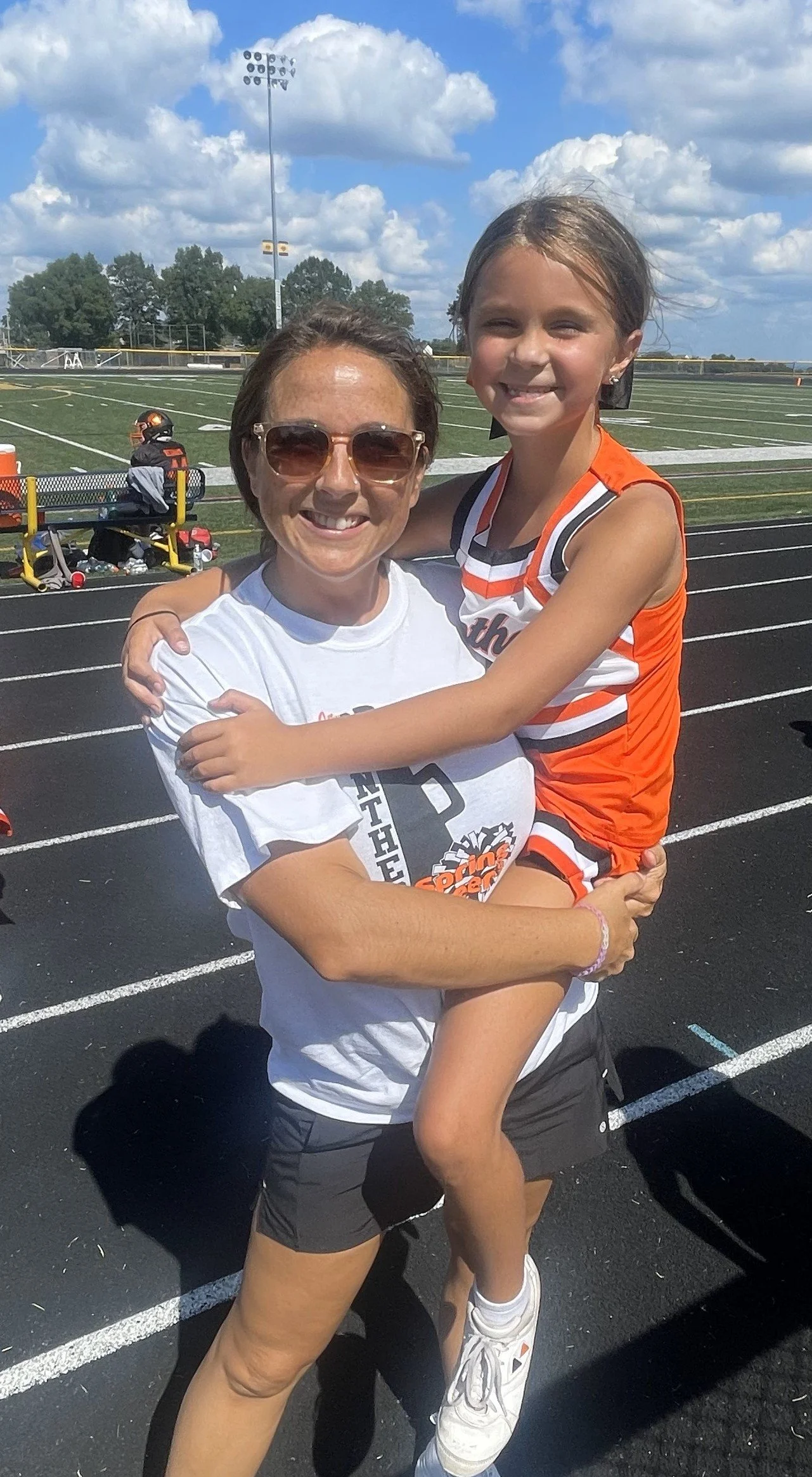 A woman holding a young girl on a running track at a sports field, both smiling. The woman has brown hair, glasses, and is wearing a white T-shirt and black shorts. The young girl is wearing an orange and white cheerleading uniform, white sneakers, and has her arms around the woman's neck. The background shows a football field, a goal post, and a partly cloudy sky.