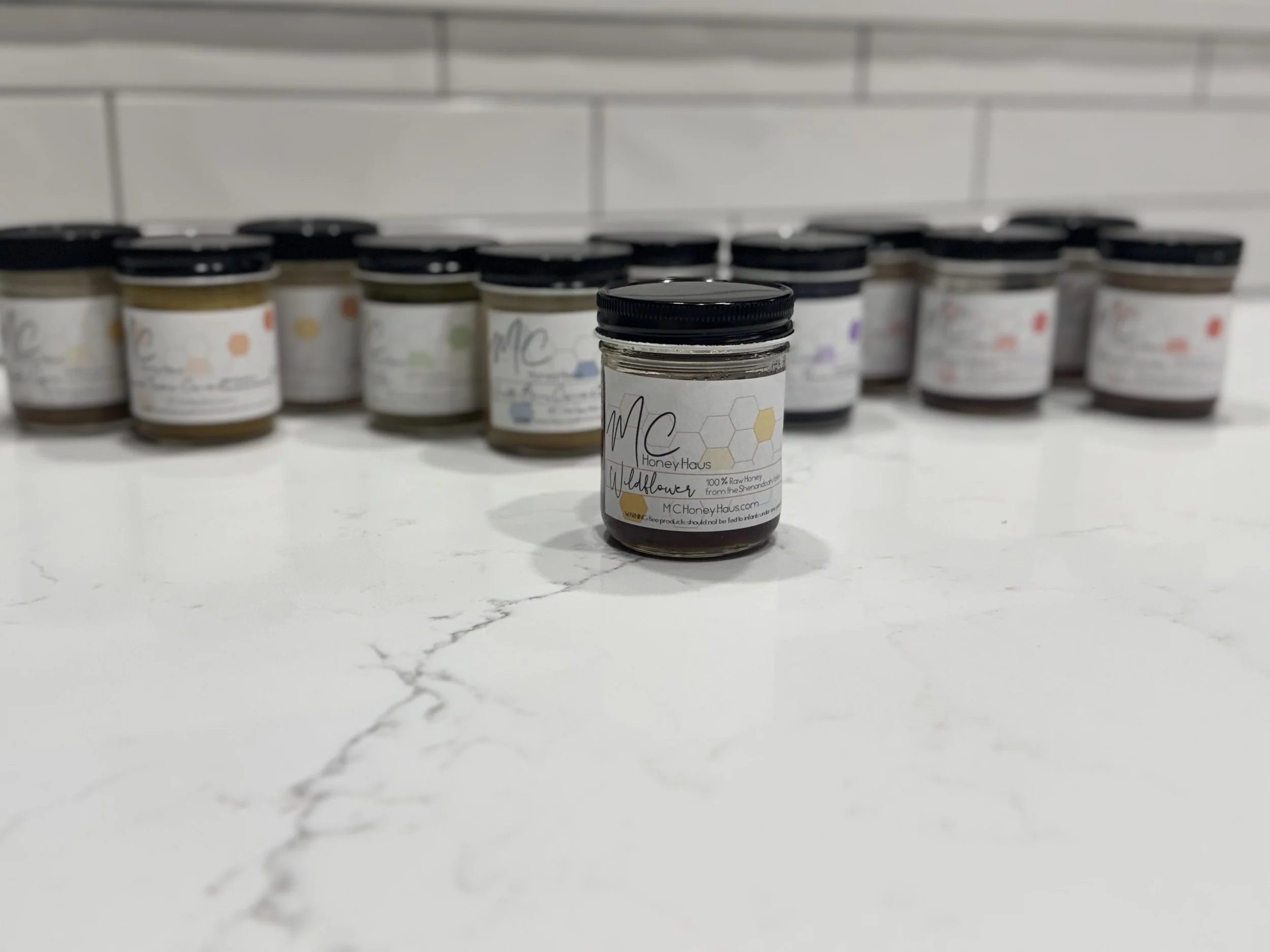 A small jar of honey in the foreground with a label from MCHoneyHaus, and other jars of honey in the background on a white marble surface.