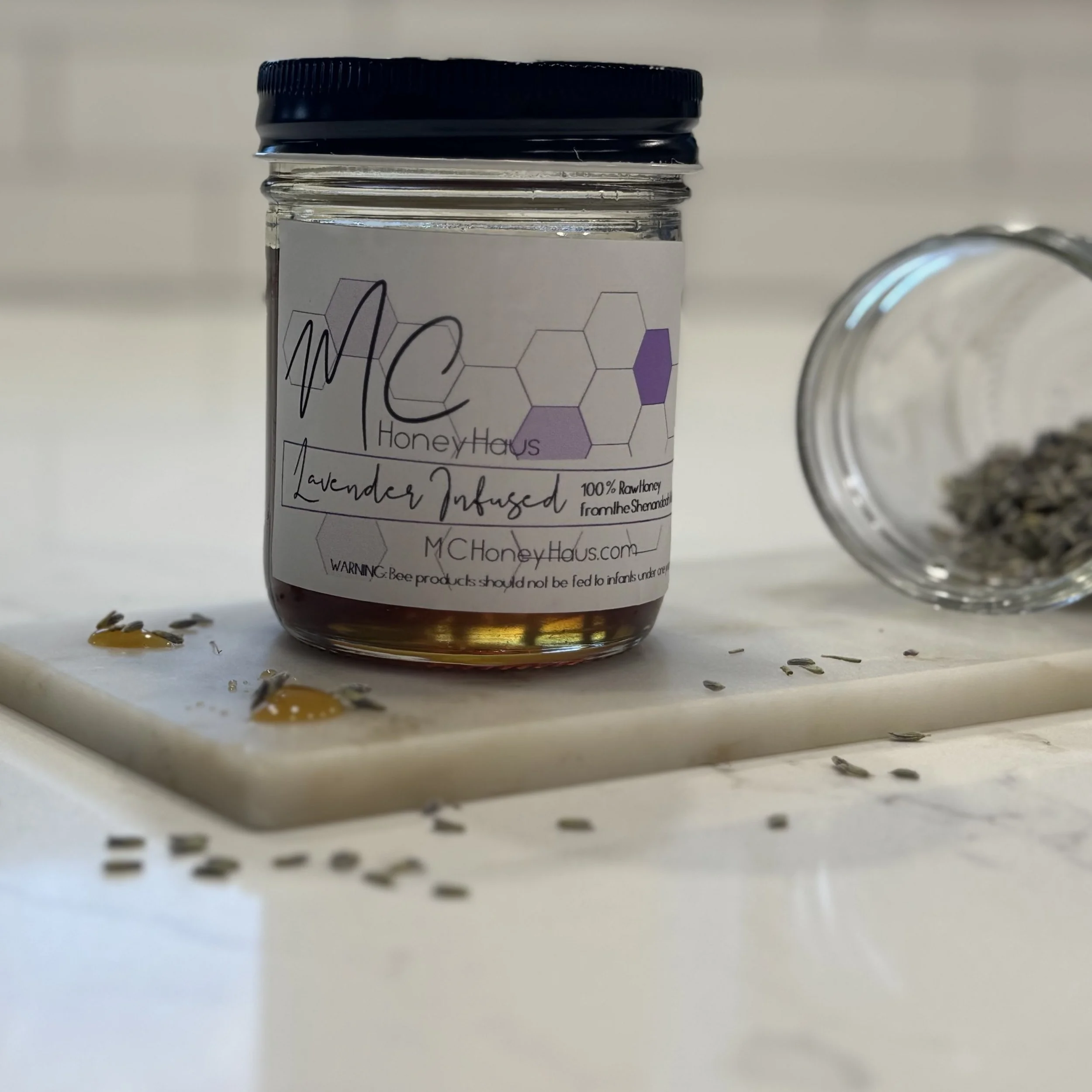 A jar of honey with a label reading 'Lavender Infused, 100% Raw Honey from the Shenandoah,' and scattered lavender seeds around it on a white surface.