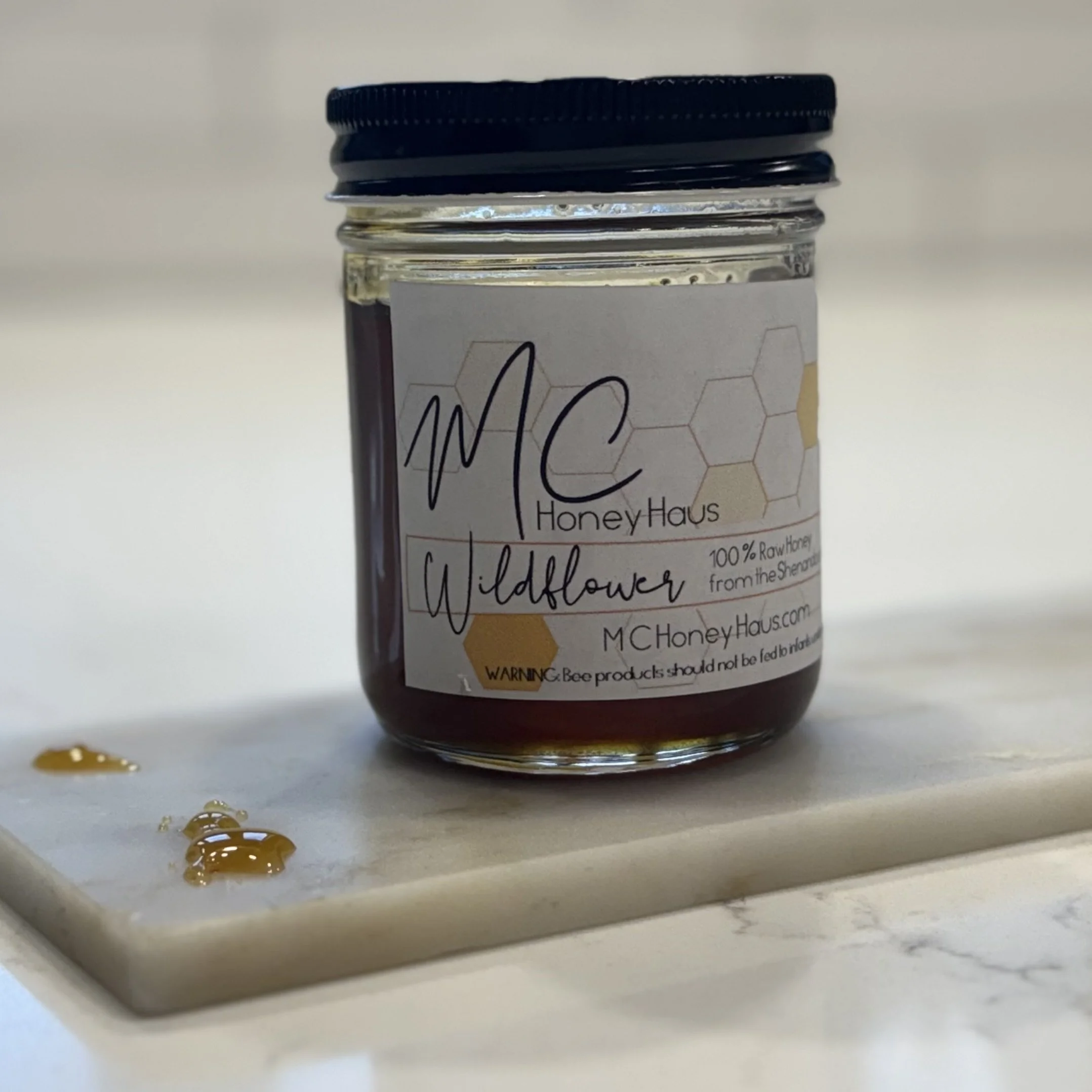 Small jar of honey labeled 'MC Honey Haus Wildflower, 100% raw honey from the Shenandoah' placed on a white surface with honey droplets nearby.