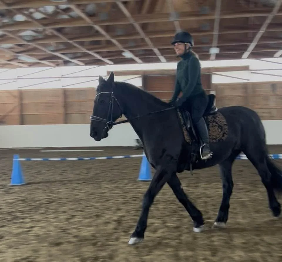 Smiling female rider wearing a helmet riding a calm black Friesian horse inside the Moose Ridge Farm arena.