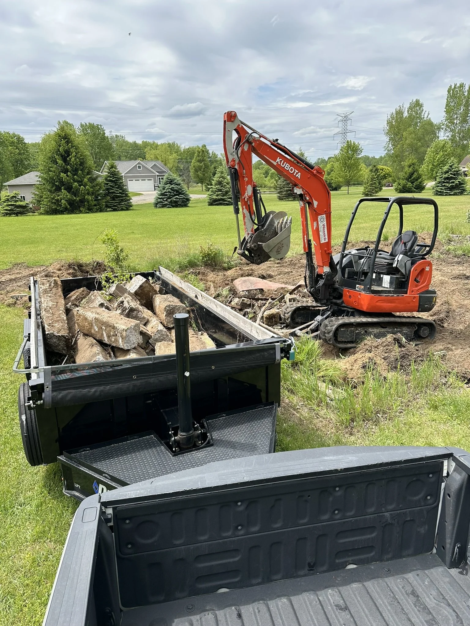 Small orange Kubota excavator digging a trench with rocks and dirt, in a green grassy area with trees and houses in the background.
