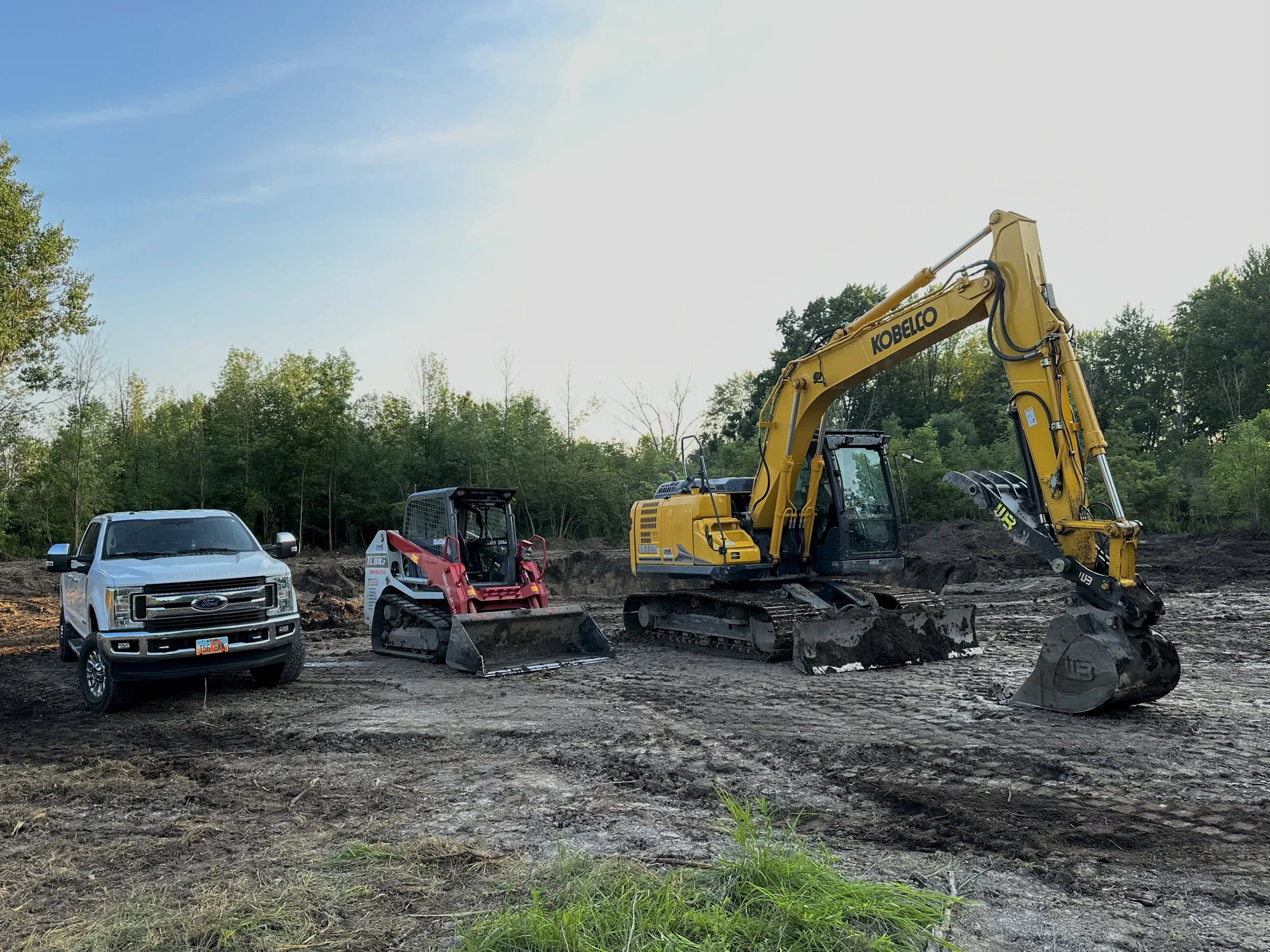 Construction site with a white pickup truck, a small red skid steer loader, and a large yellow excavator on cleared ground with trees in the background.