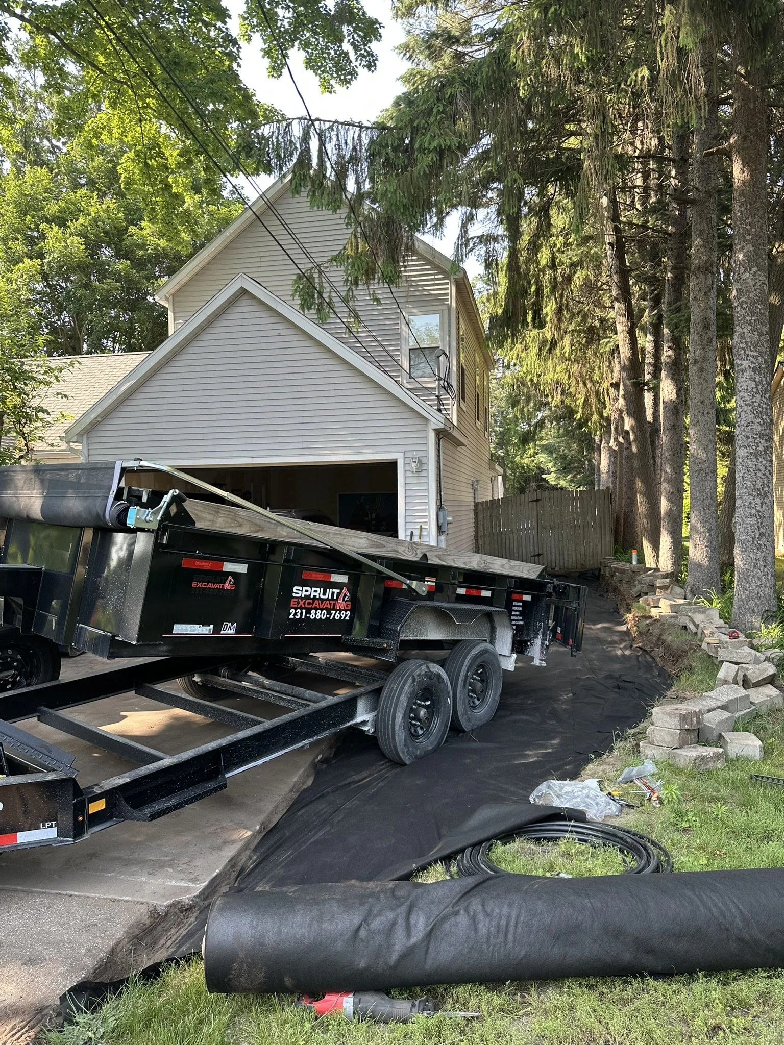 A house with a garage surrounded by trees, a trailer parked on a driveway, and black fabric and construction materials on the ground.