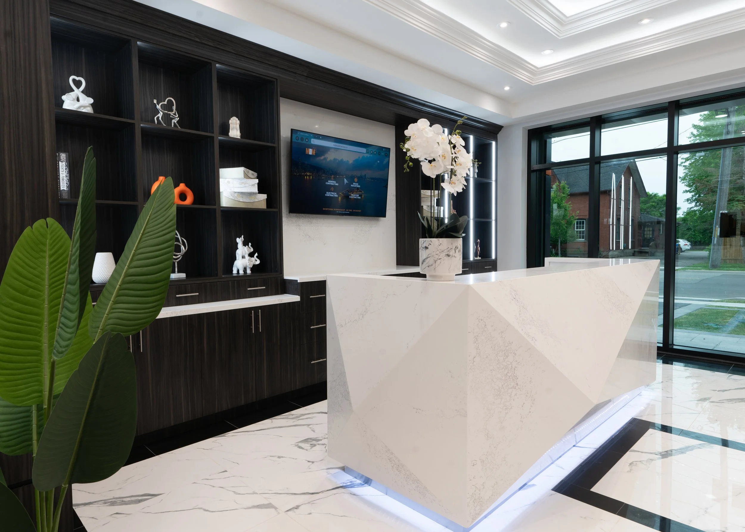 Modern hotel reception area with a white geometric reception desk, a tall vase with white orchids on top, dark wood shelving with decorative items, large windows showing a street view, and marble flooring.
