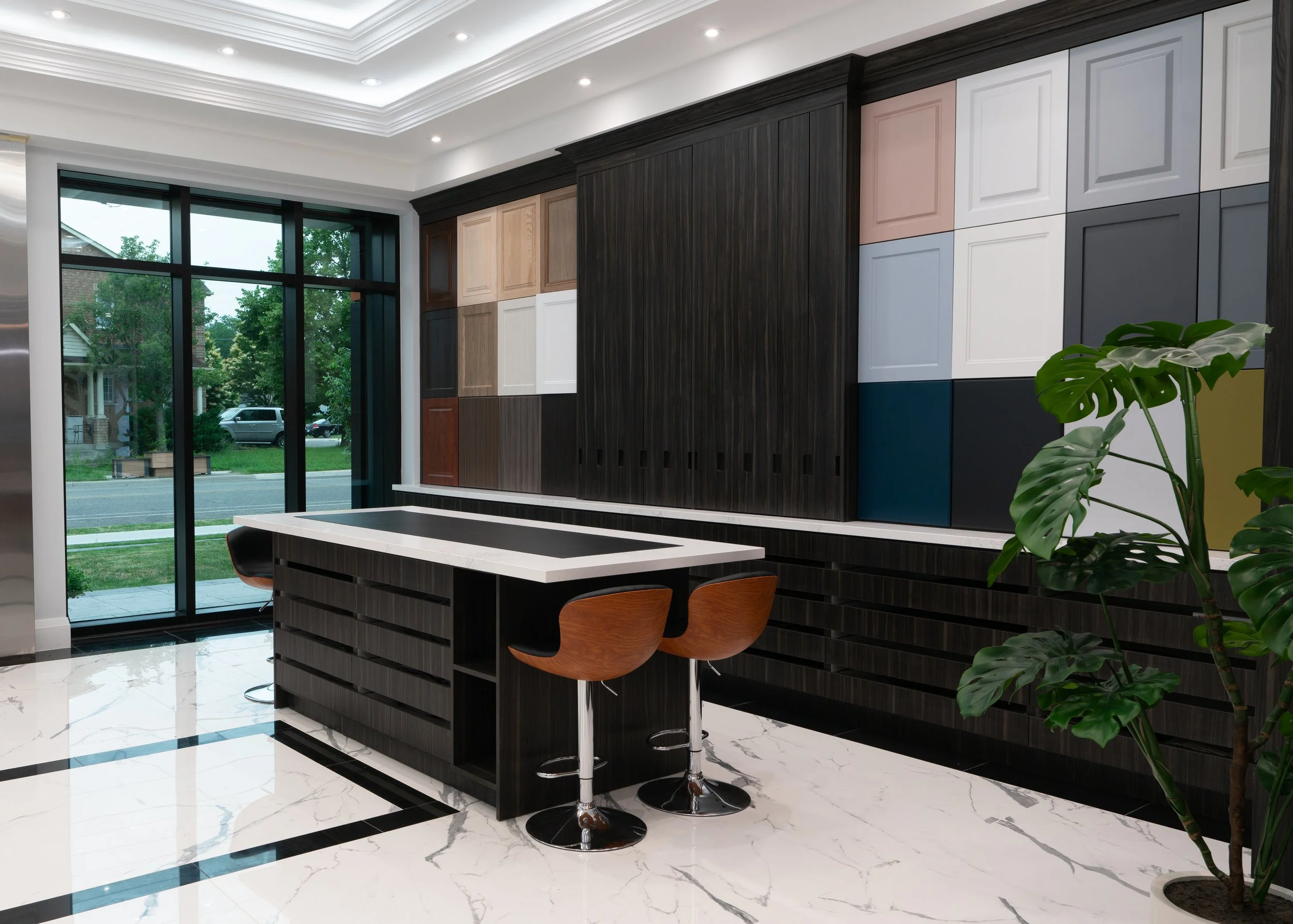 Modern kitchen with black cabinetry, colorful square cabinet doors on the wall, a white marble countertop island, two brown bar stools, a large window with street view, and a green potted plant.