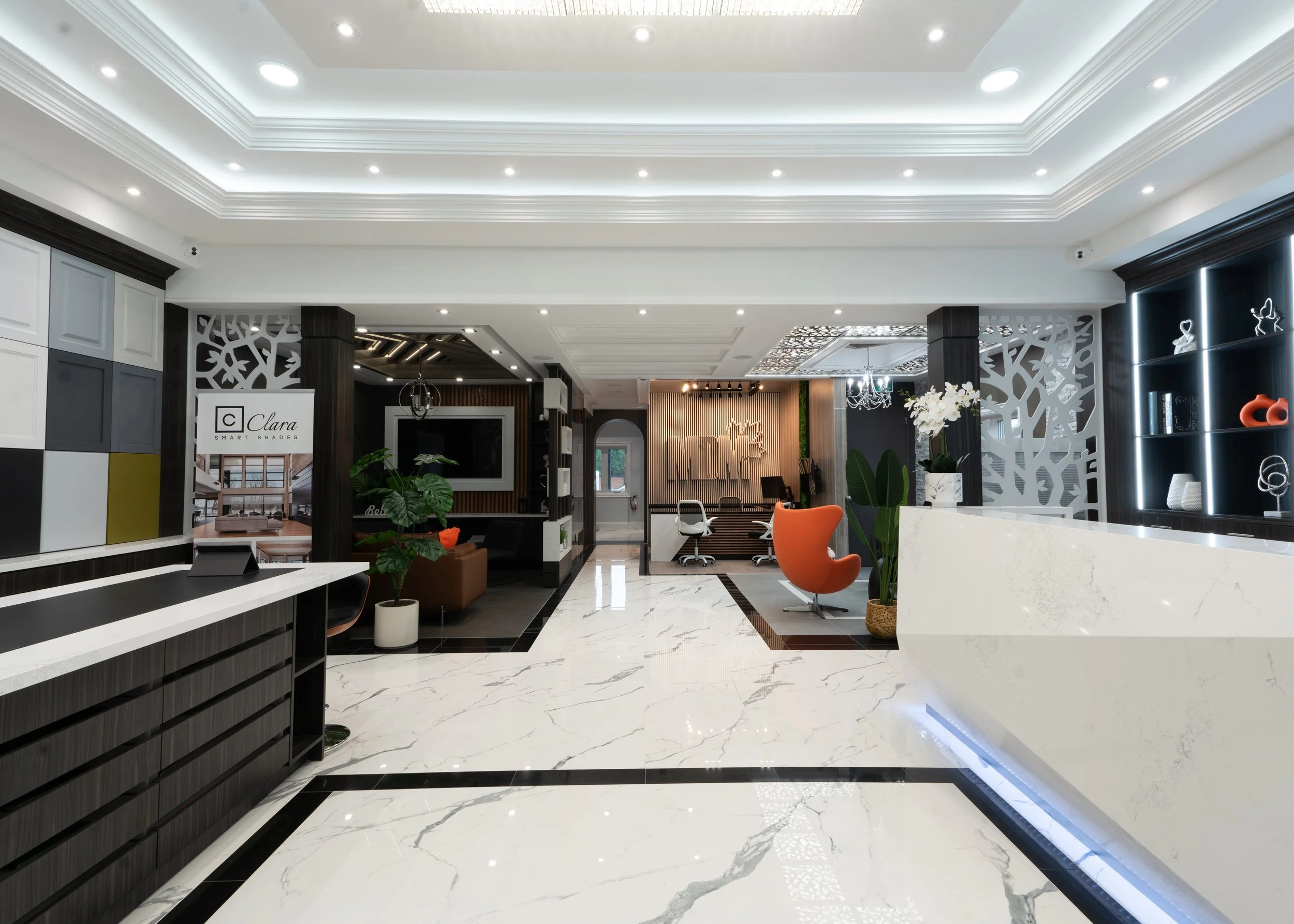 Modern interior of a stylish reception or lobby area with white marble floors, black and white walls, decorative lighting, green plants, and contemporary furniture including orange chairs and a white reception desk.