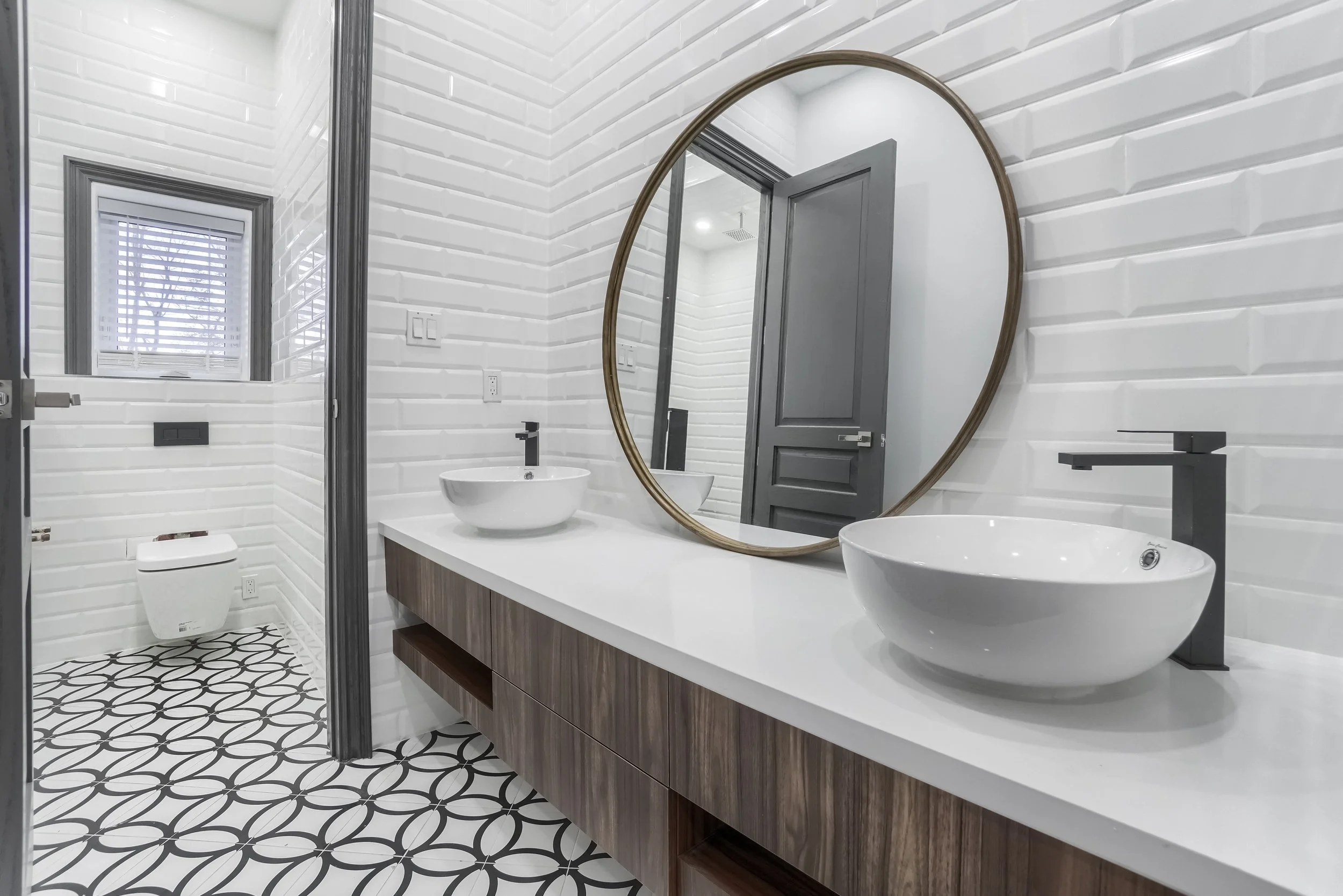 Modern bathroom with white brick walls, a large round mirror, two vessel sinks, black faucets, a wooden vanity, a small window with blinds, a toilet, and black and white patterned floor tiles.