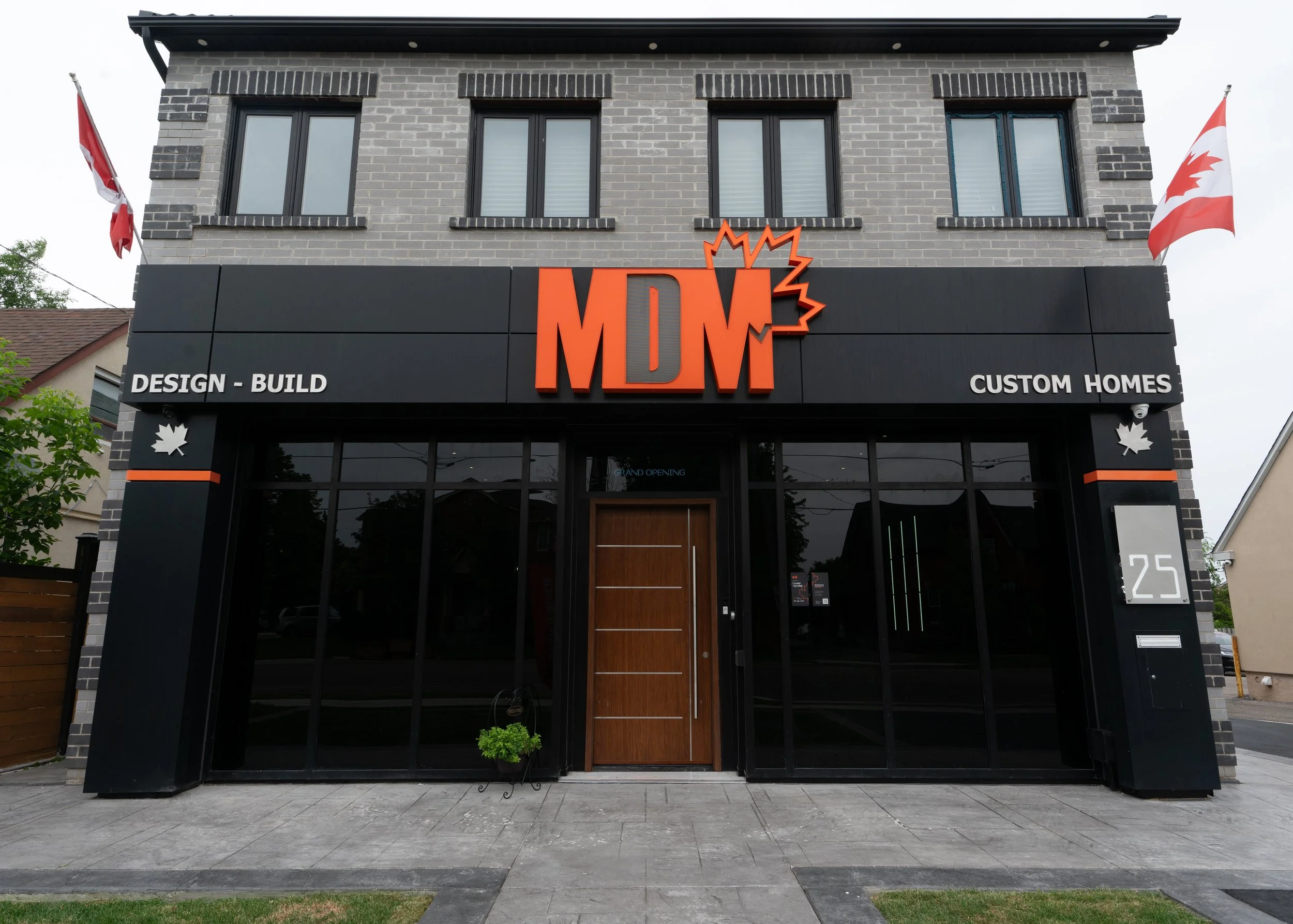 Facade of a black and gray brick commercial building with large glass windows, a central wooden door, and an orange and black MDW sign on the upper black section. Signs on either side advertise design-build and custom homes. Canadian flags are on eit