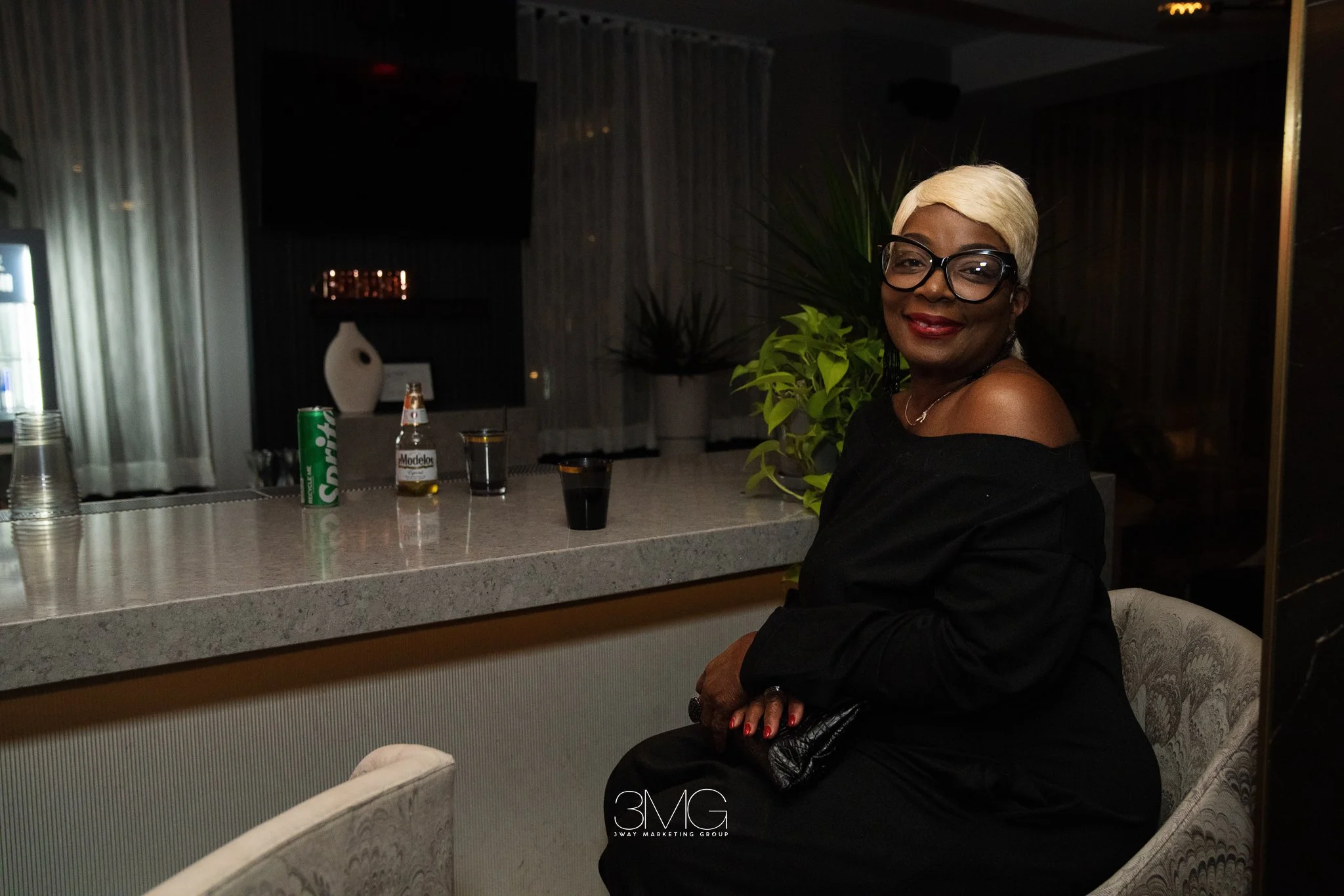 A woman with short platinum blonde hair, black glasses, and red lipstick sitting at a bar counter in a dimly lit lounge or bar, smiling. There are drinks, including a green can and a bottle of Modelo beer, on the counter. Behind her, there is a plant