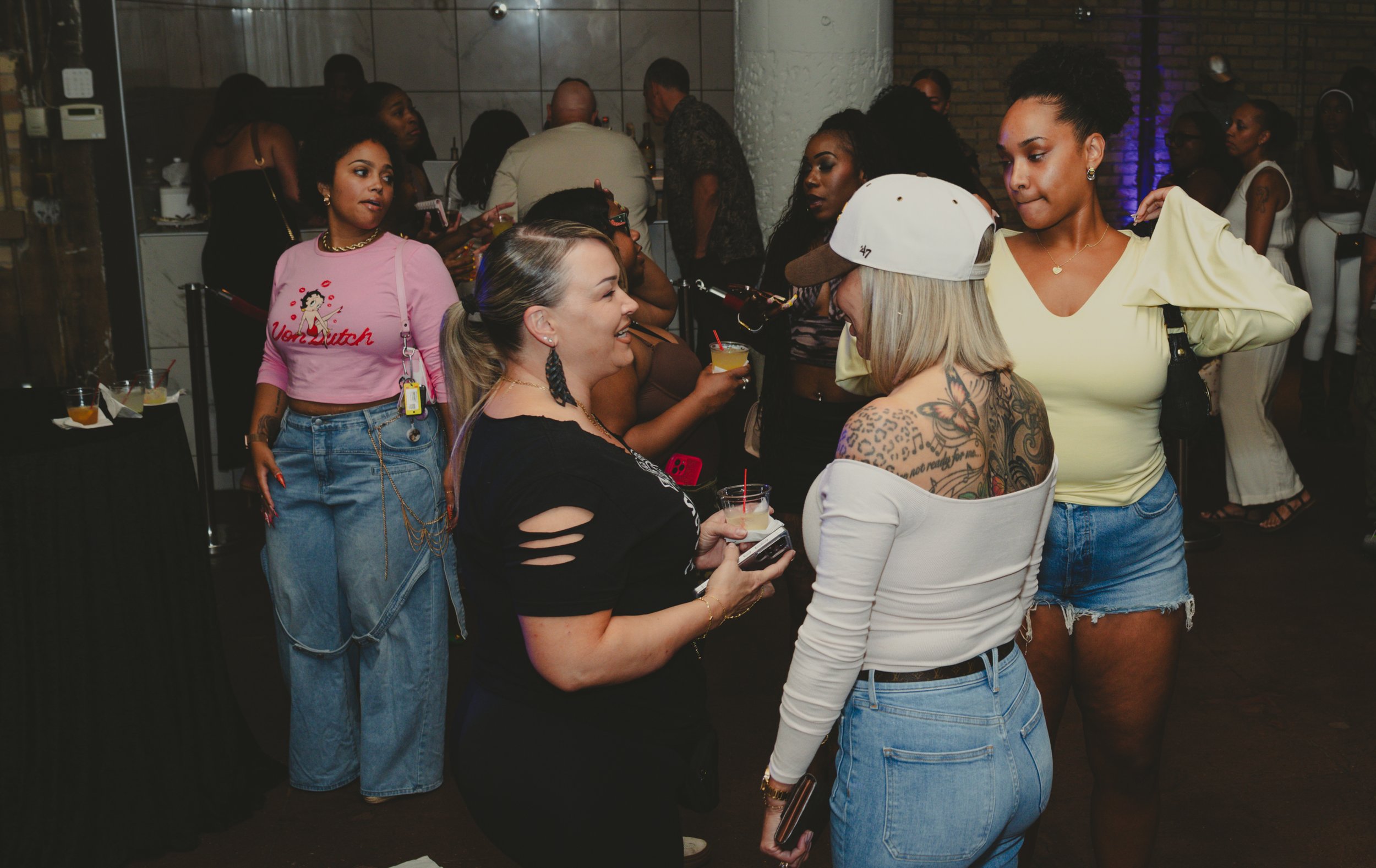 Group of women and one man socializing, holding drinks at an indoor party or event with a lively atmosphere.
