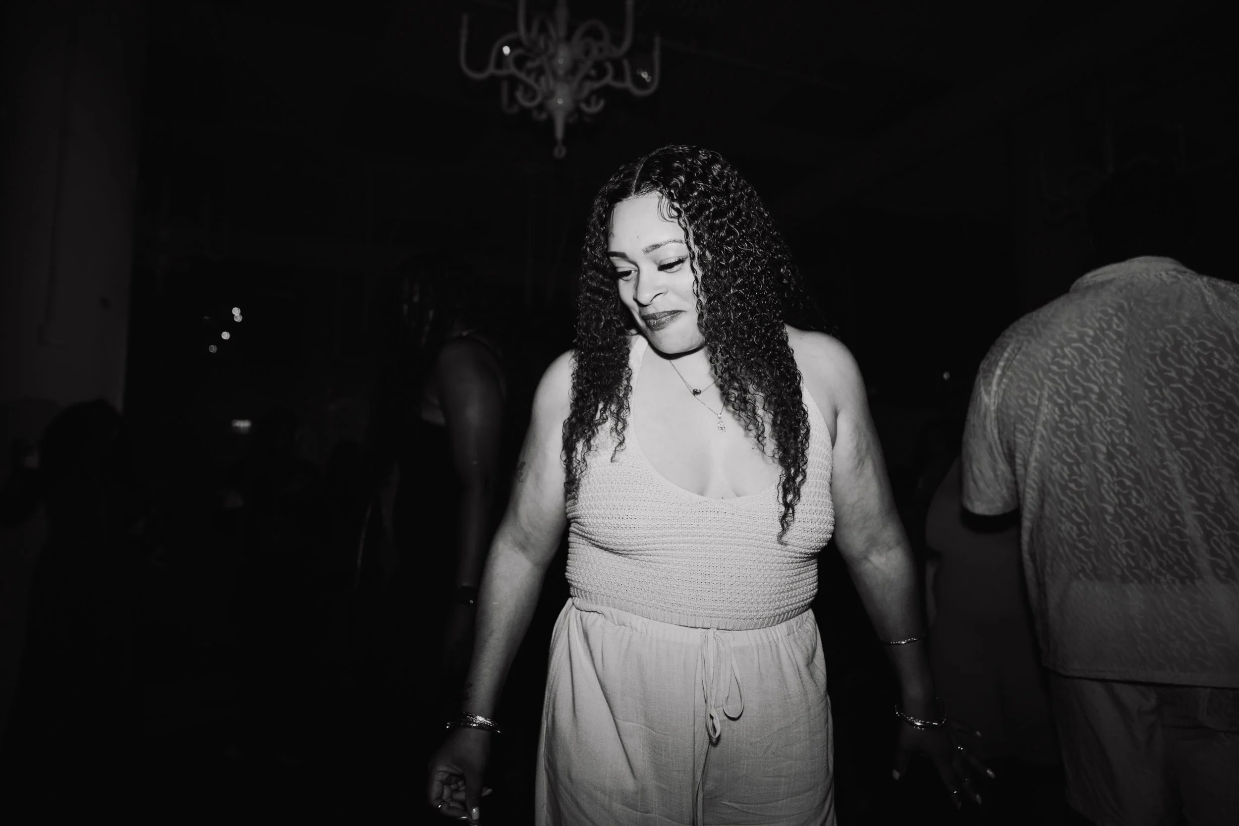 A woman with curly hair wearing a light-colored tank top and light-colored pants, standing in a dark environment, possibly at a party or nightclub, with other people partially visible around her.