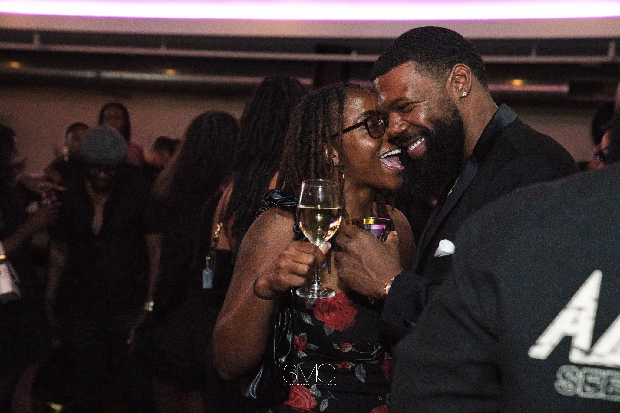 A smiling woman with glasses and dreadlocks holding a glass of champagne, leaning into a man with a beard and an earring, both smiling and dancing at a lively gathering or party.