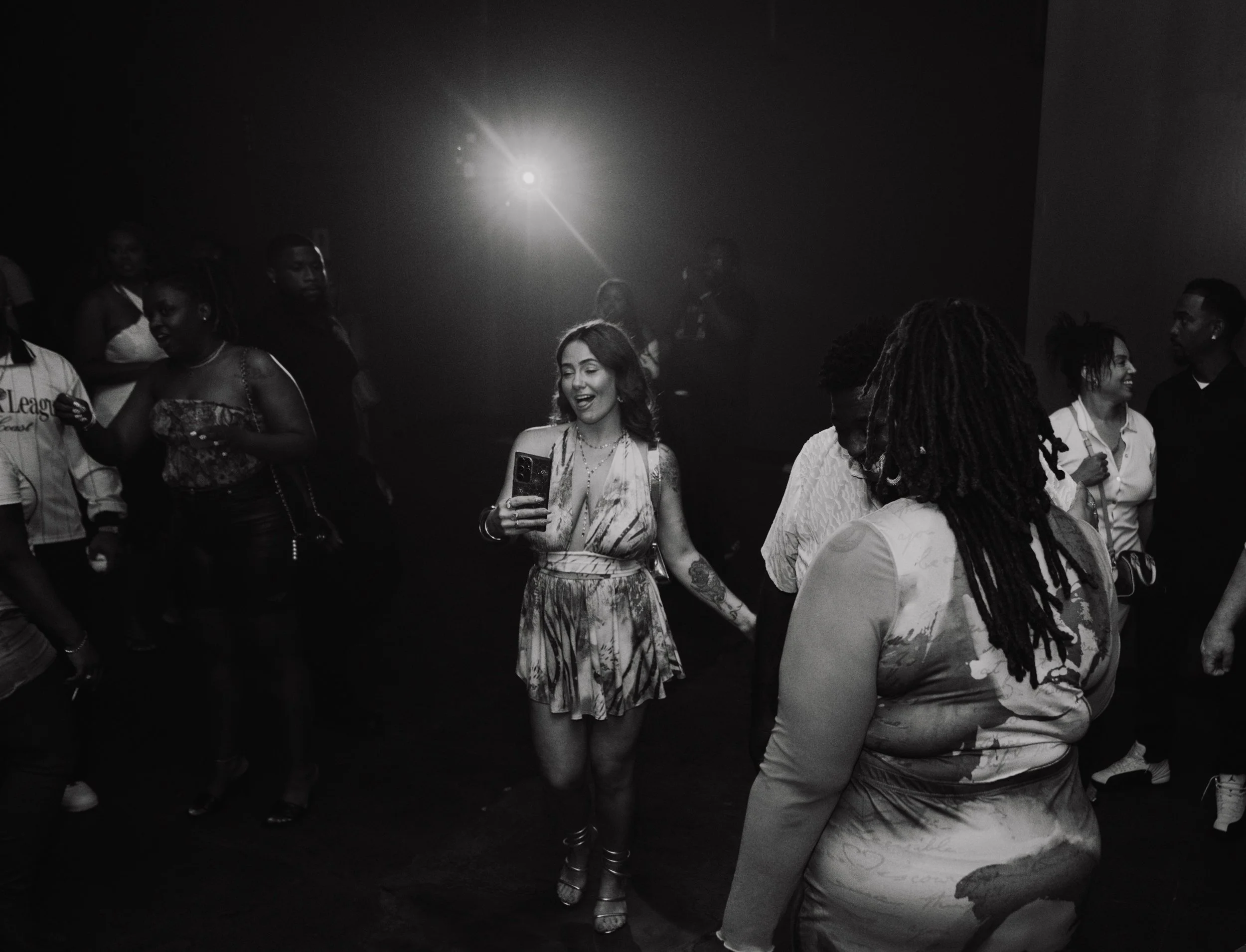 People standing and talking at what appears to be a social gathering or event, with some individuals smiling and looking at their phones, in black and white.