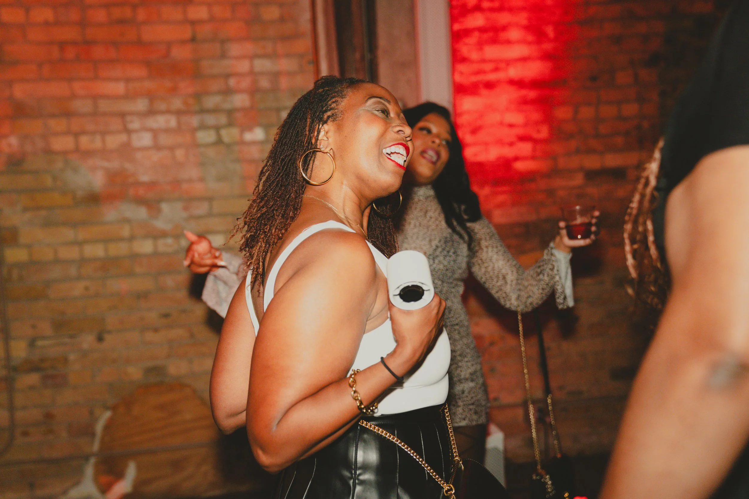 A woman with braided hair, hoop earrings, and a white top, smiling and holding a drink at a social gathering with a brick wall background and red lighting.