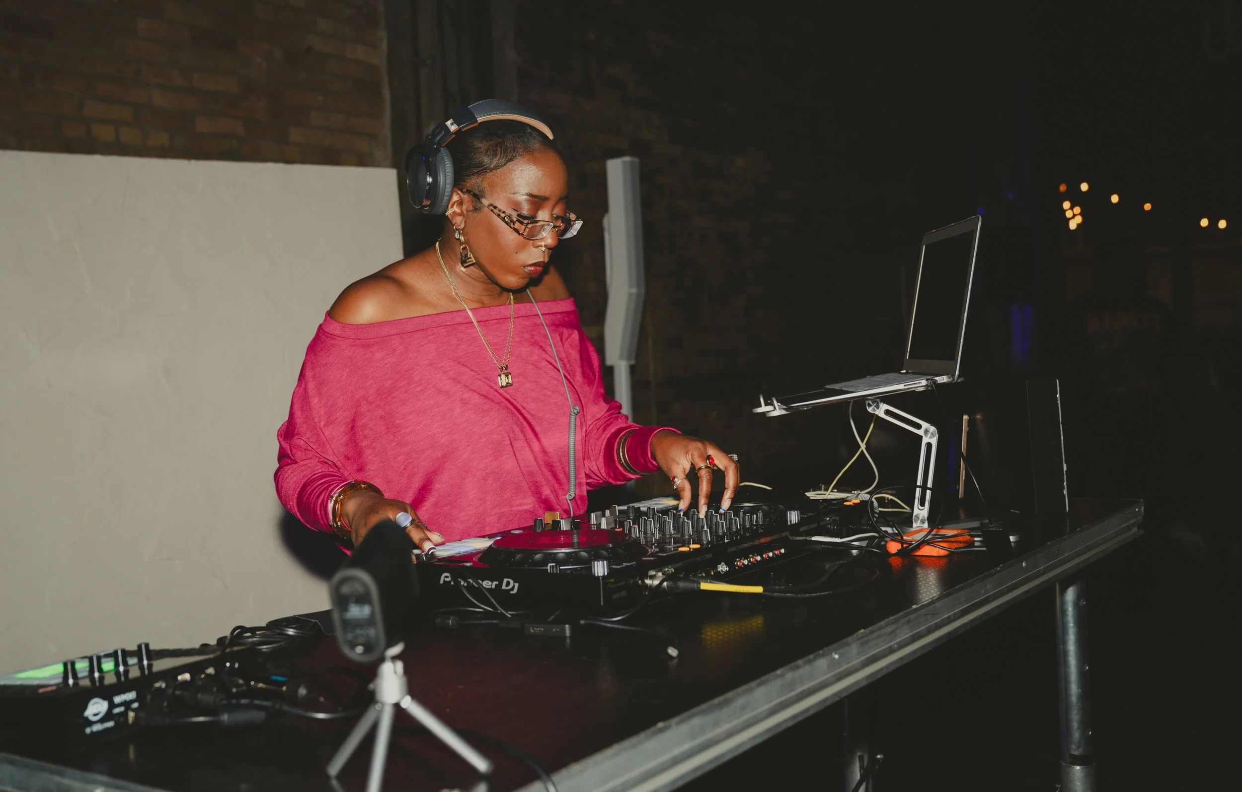 A woman DJ wearing glasses and colorful jewelry, in a pink off-shoulder top, DJing with headphones at a nightclub or event at night.