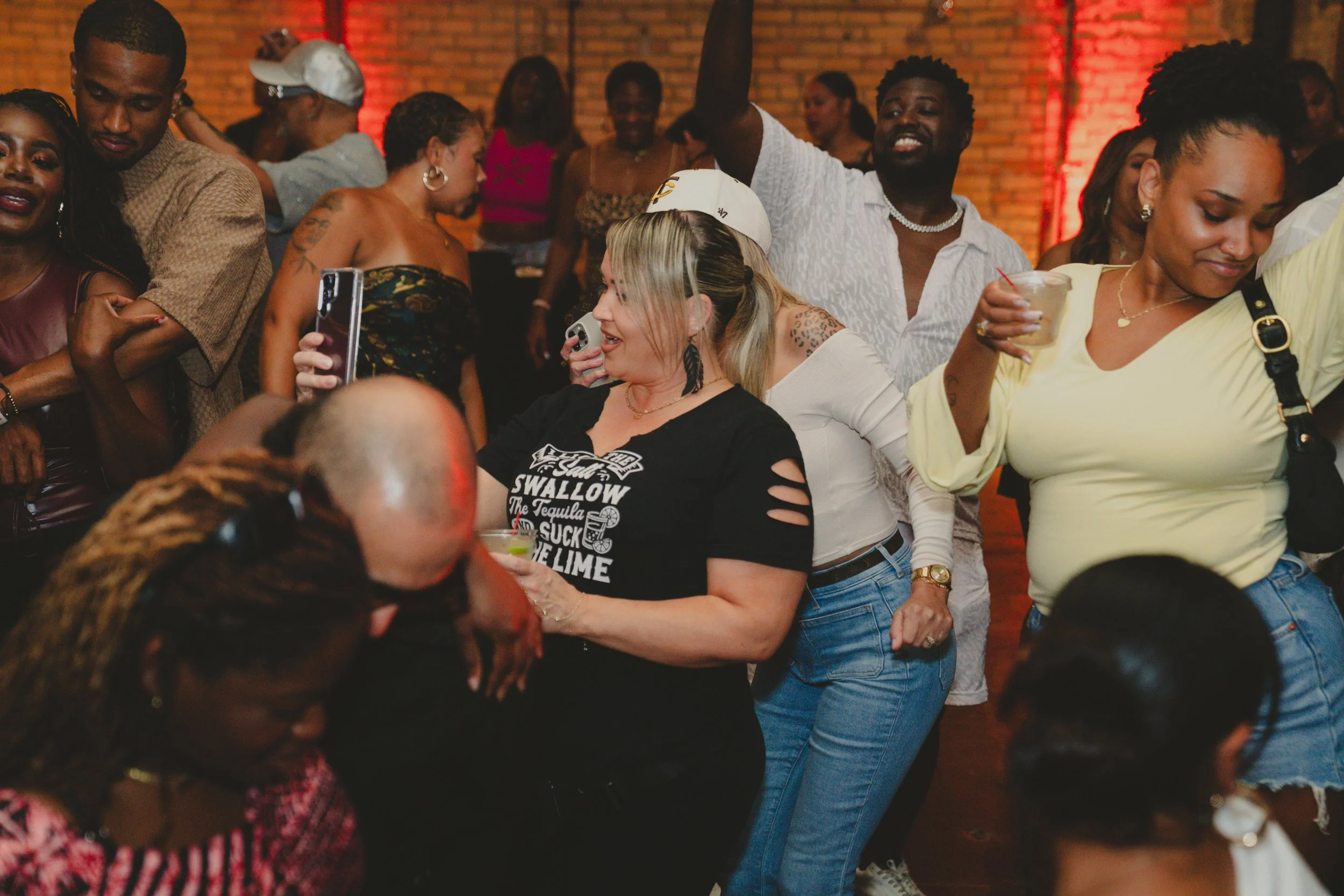 People dancing and socializing at a party with brick walls and red lighting.