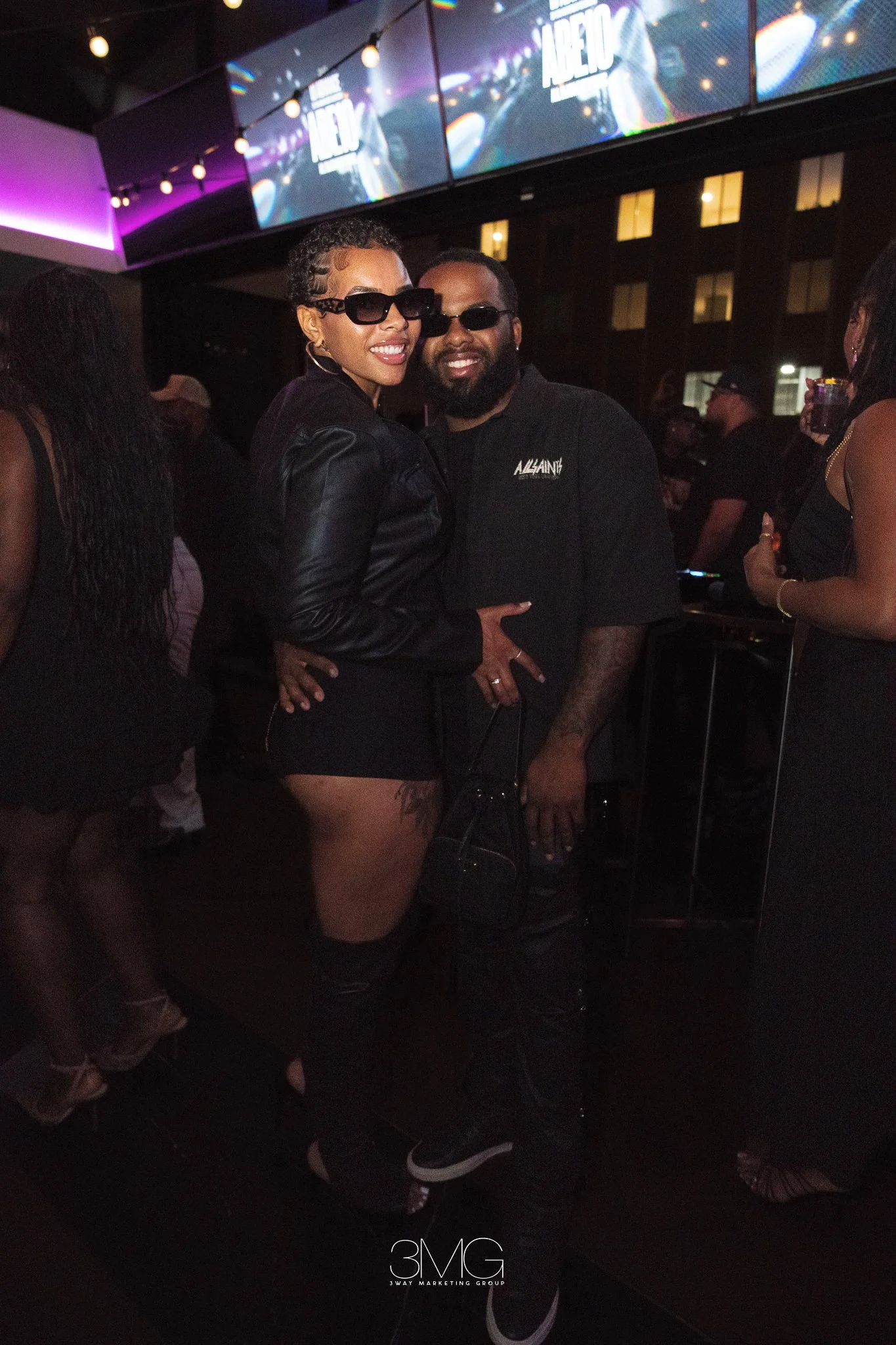 A woman and a man in sunglasses smiling and posing together at a nightclub or party. The woman is wearing a black leather jacket and shorts, and the man is wearing a black t-shirt with white text and glasses. There are other people in the background,