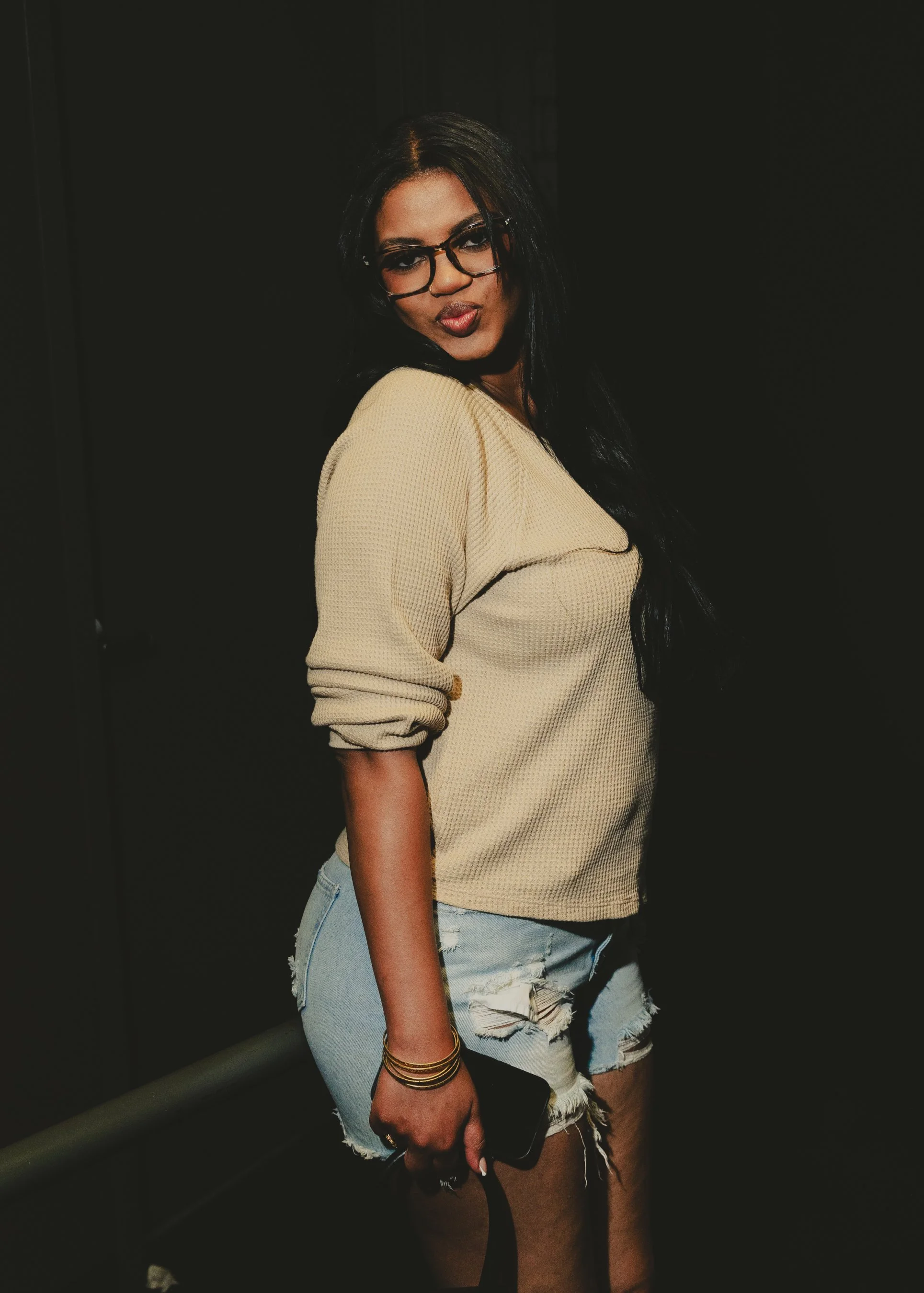 A woman with long black hair, glasses, wearing a beige sweater and ripped denim shorts, holding a phone and standing against a dark background.