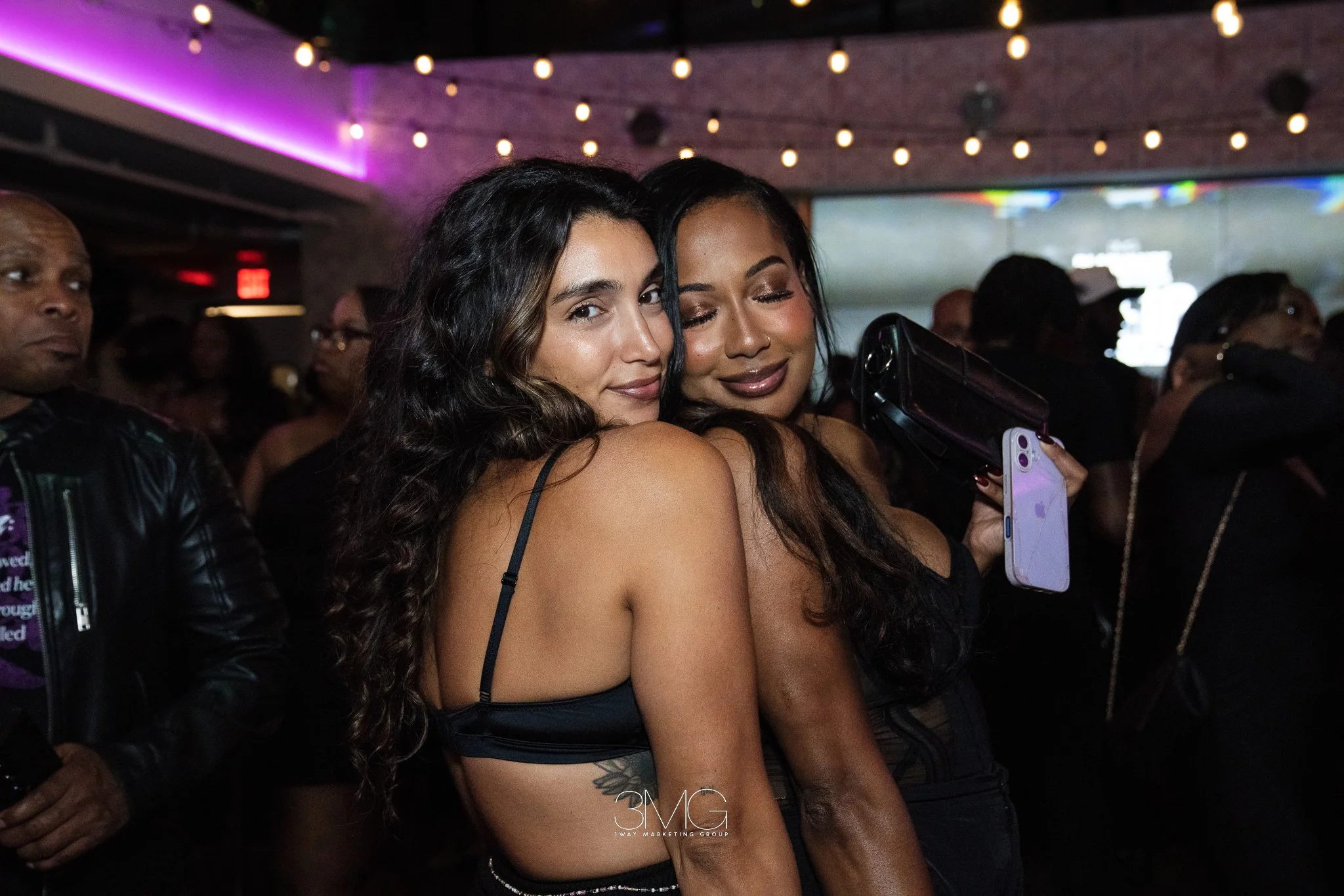 Two women with long wavy hair taking a selfie together at a dimly lit indoor event with string lights overhead. One woman has her eyes closed and is smiling while the other woman has her eyes open, smiling, and holding a phone.