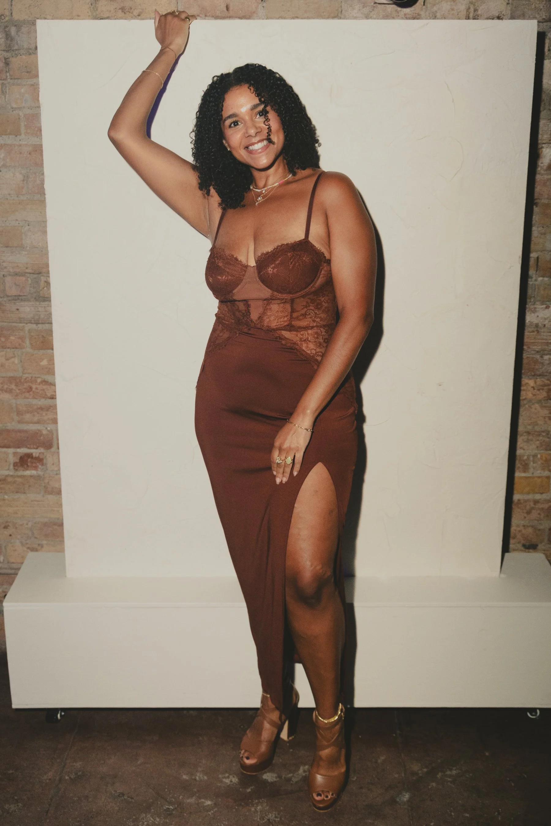 A woman with curly black hair wearing a brown lace dress and high heels, smiling and posing with her hand raised, standing in front of a white backdrop with brick wall to the side.