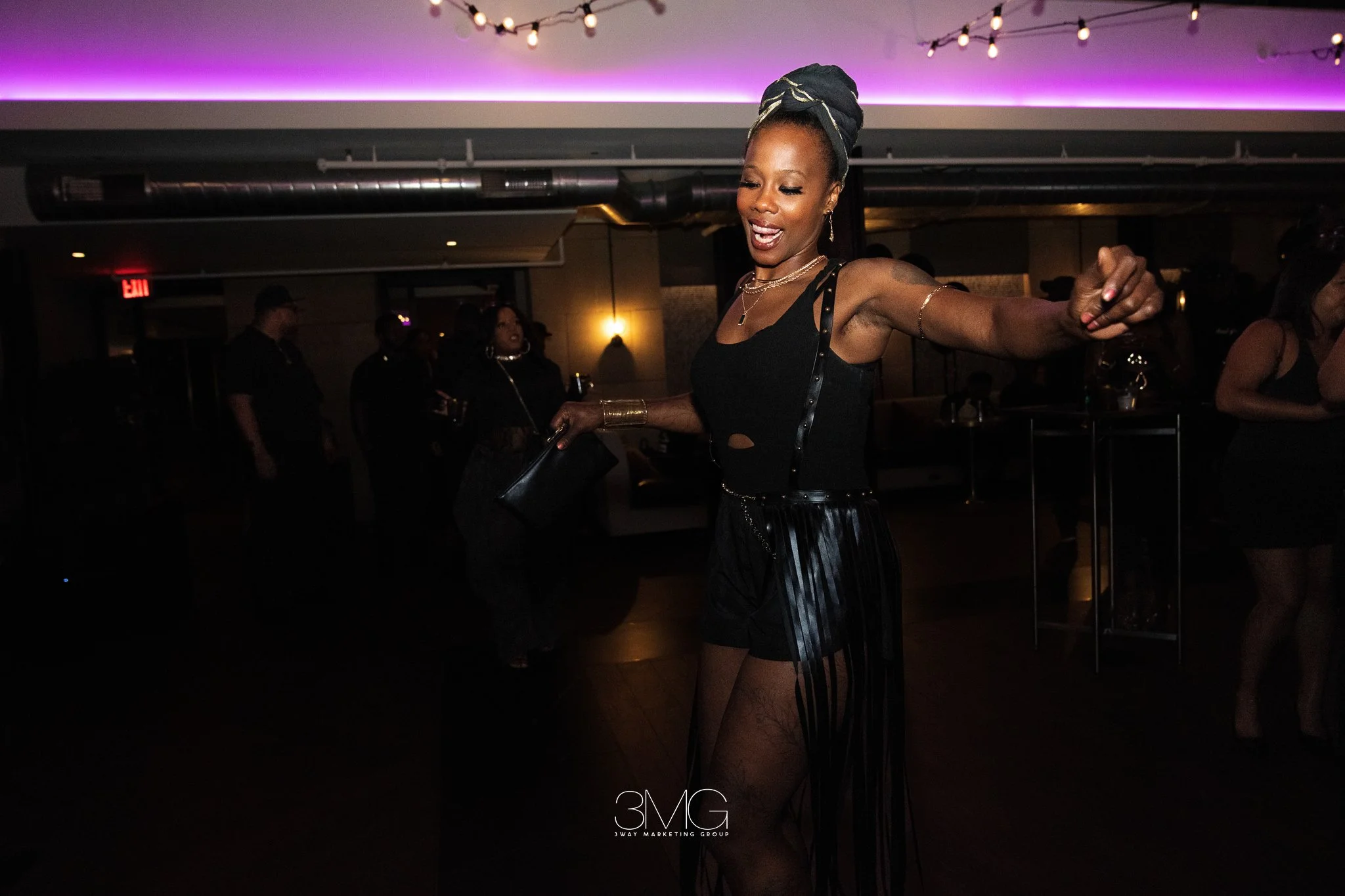 A woman dancing at a party or club with dark lighting, dressed in black with a head wrap, jewelry, and a sheer skirt. People are in the background, and there are purple and yellow lights.