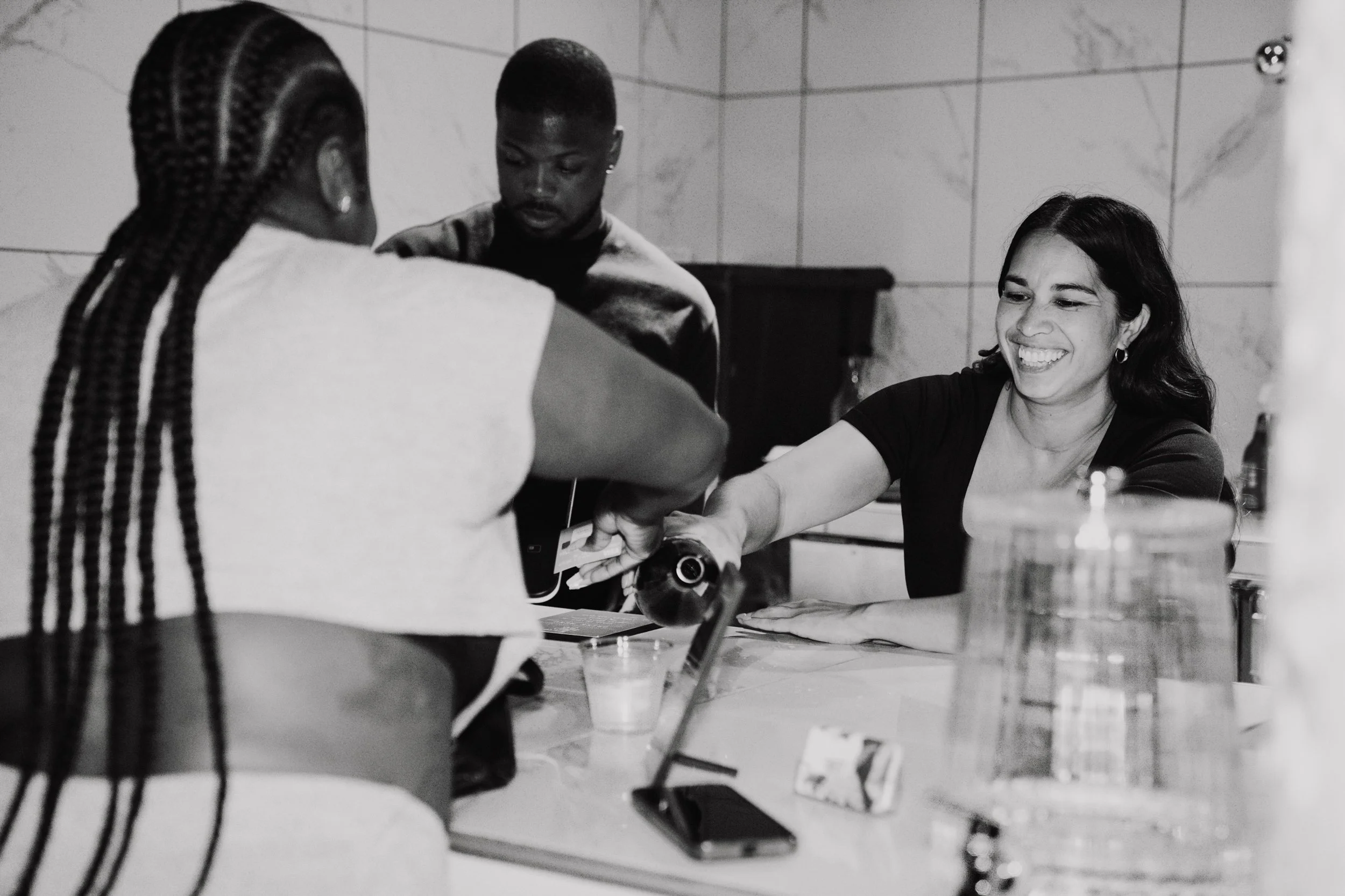 Three women gathered around a table, engaged in a conversation or activity, with one smiling, in a room with tiled walls.