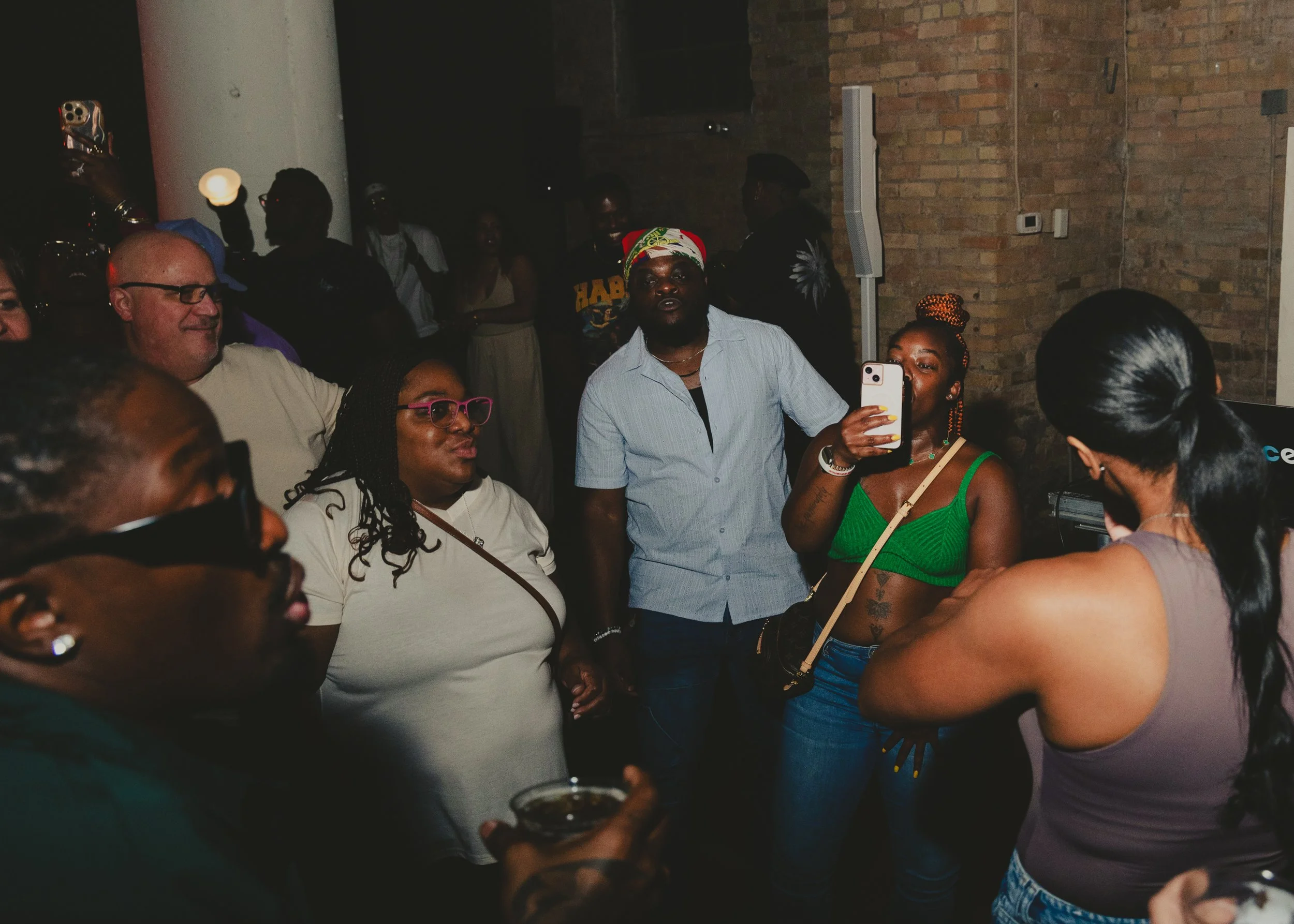Group of diverse people socializing at a party in a dimly lit indoor venue with brick walls. Some people are taking photos, while others are talking and enjoying drinks.