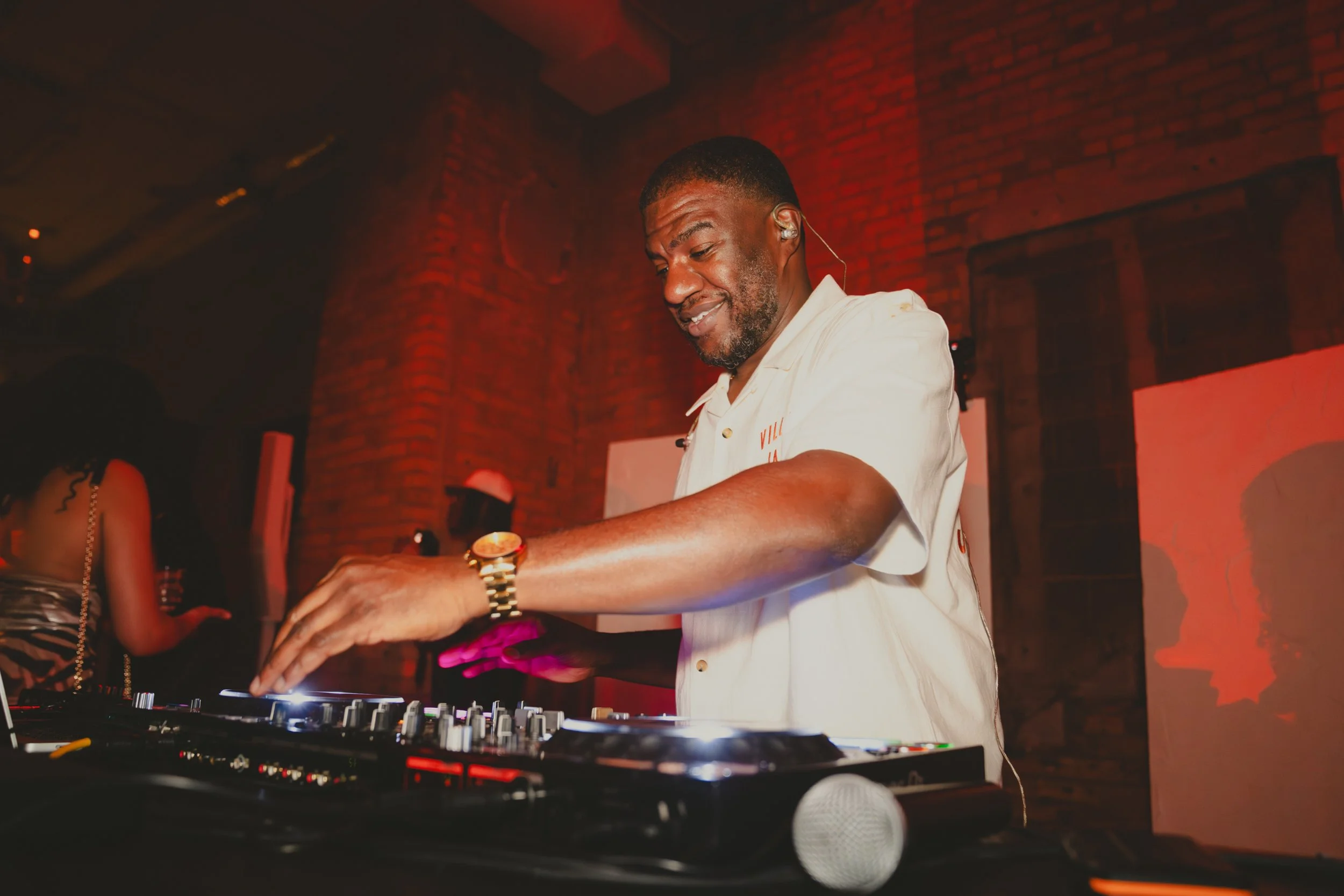 A man DJing at a nightclub with a brick wall background, wearing a white shirt, gold watch, and in-ear monitor.