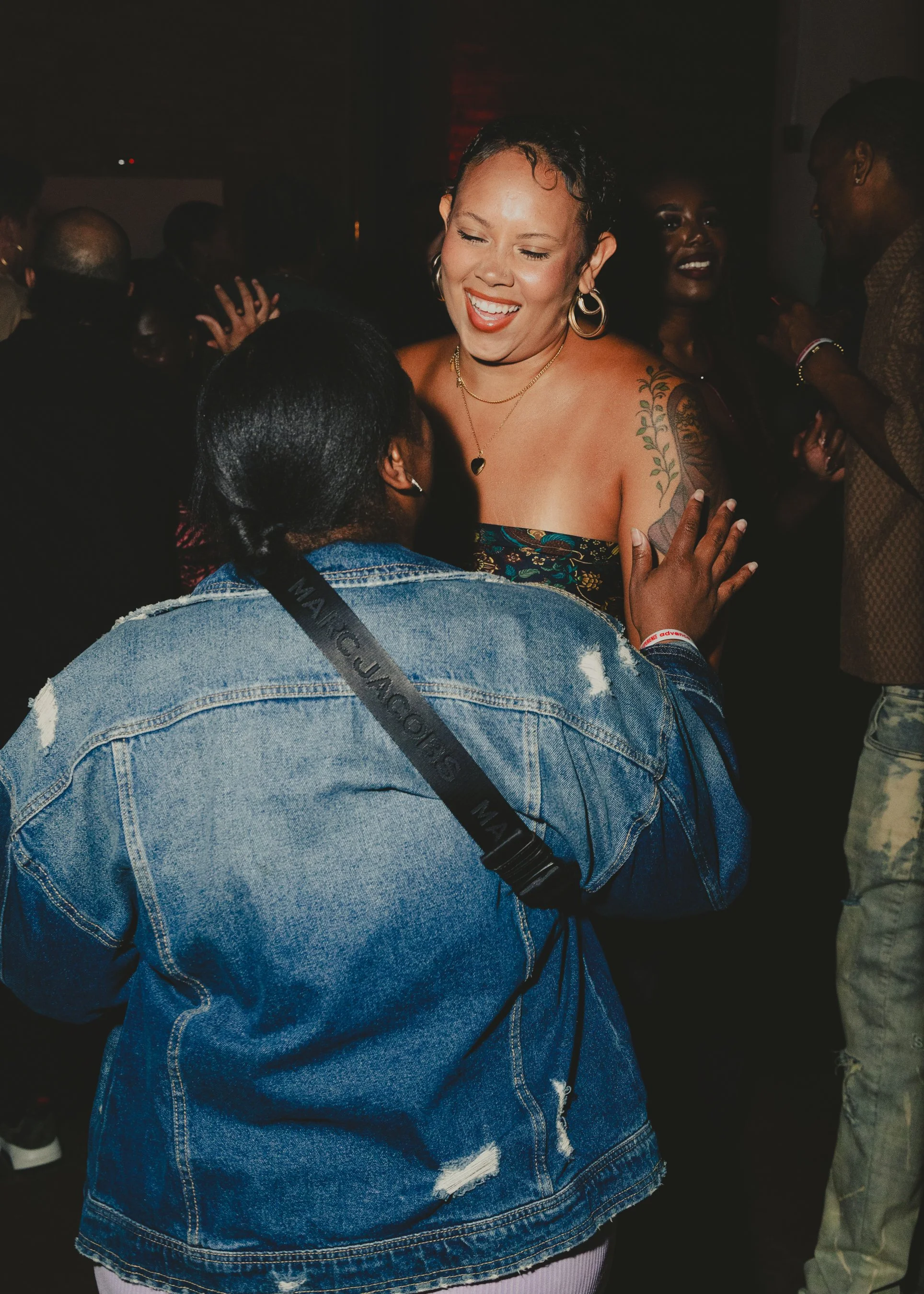 A woman with short hair, earrings, and tattoos on her shoulder, smiling and dancing at a crowded indoor event while talking to another person with braided hair.