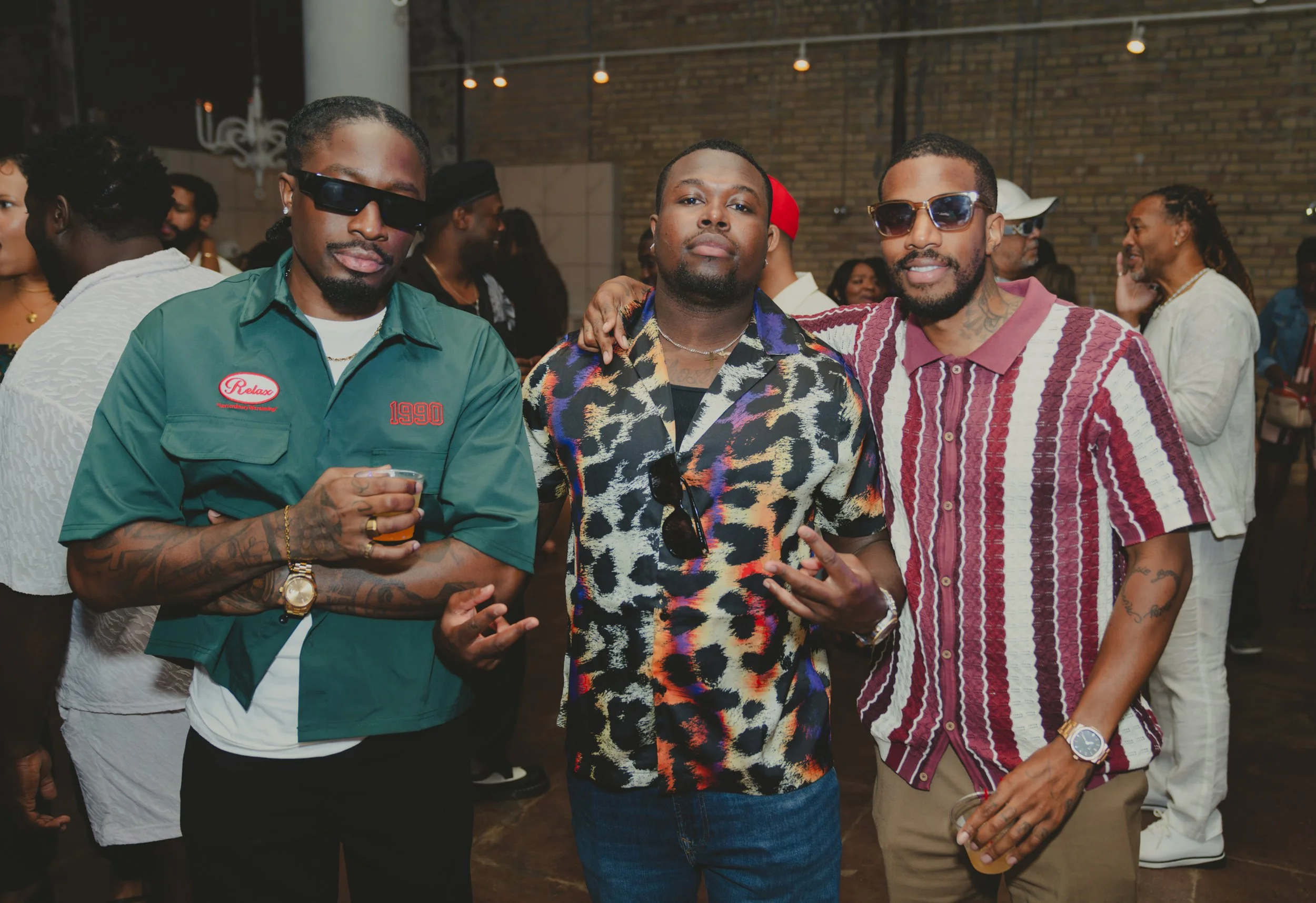 Three men posing together at a social event, with other people in the background, in a room with brick walls and warm lighting.