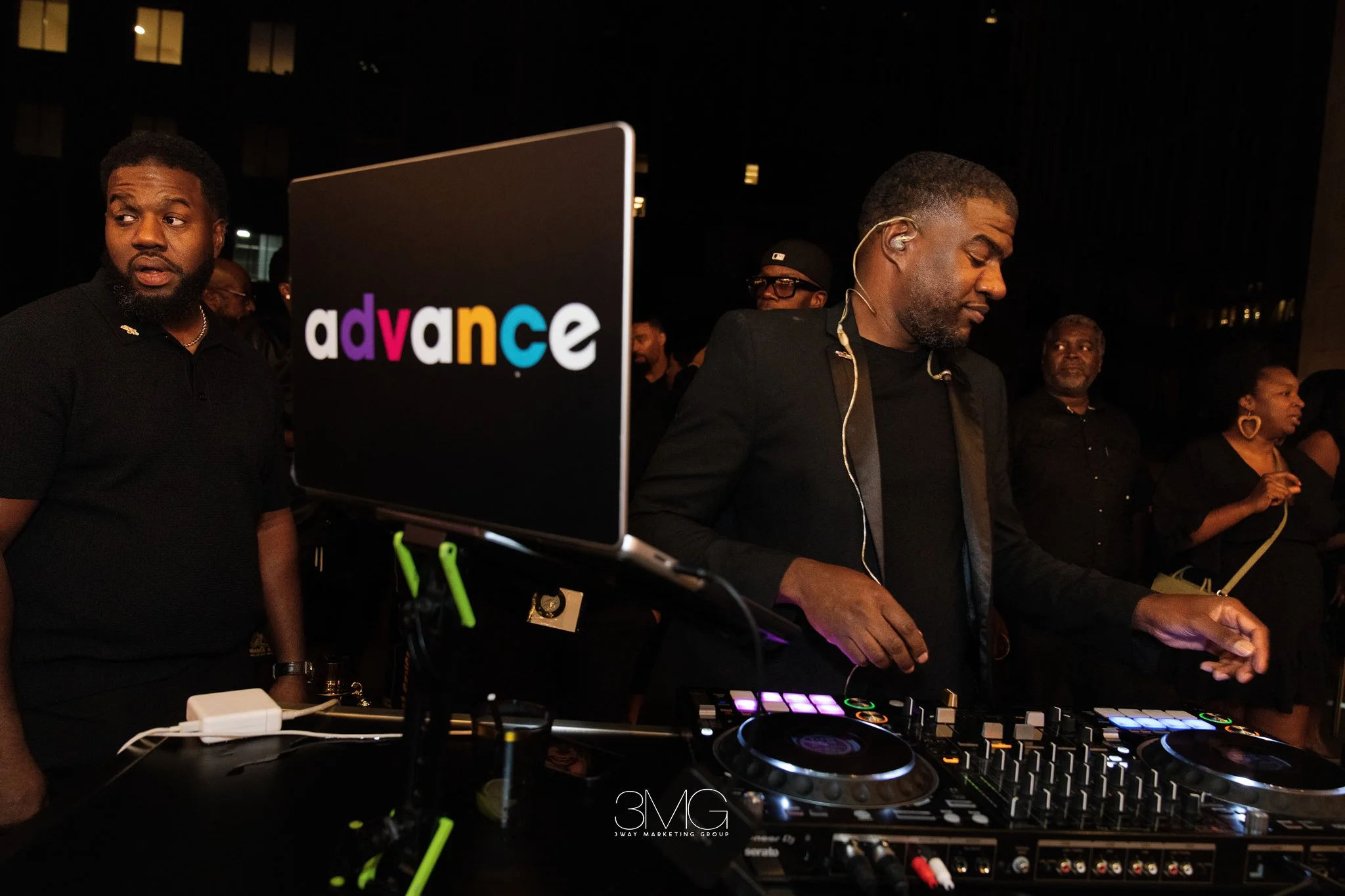 A DJ booth at an event with people around, with a focus on a DJ operating equipment. One person appears to be DJing with a turntable and mixer. The background is dark, suggesting an indoor venue at night. The screen in the foreground displays the log