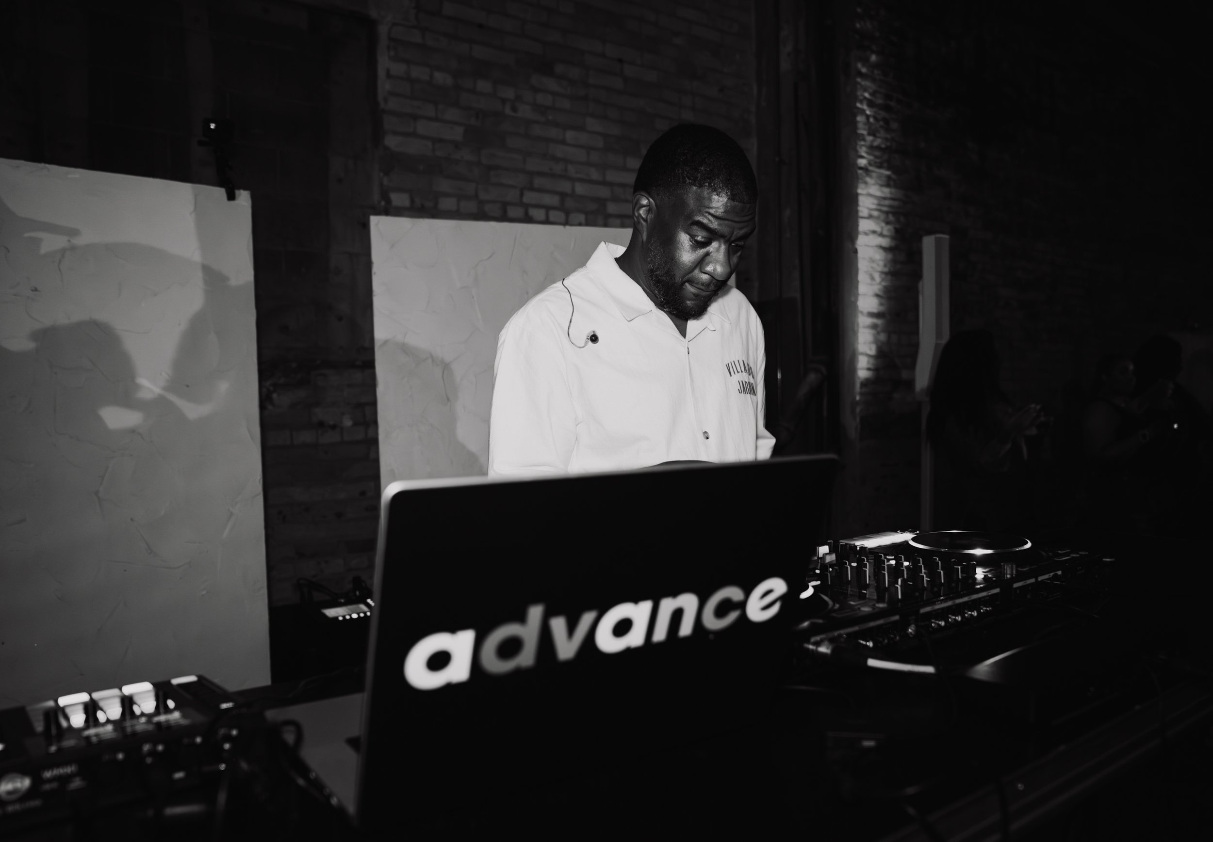 A man in a white shirt standing behind DJ equipment at a music event, with a dark brick wall in the background.