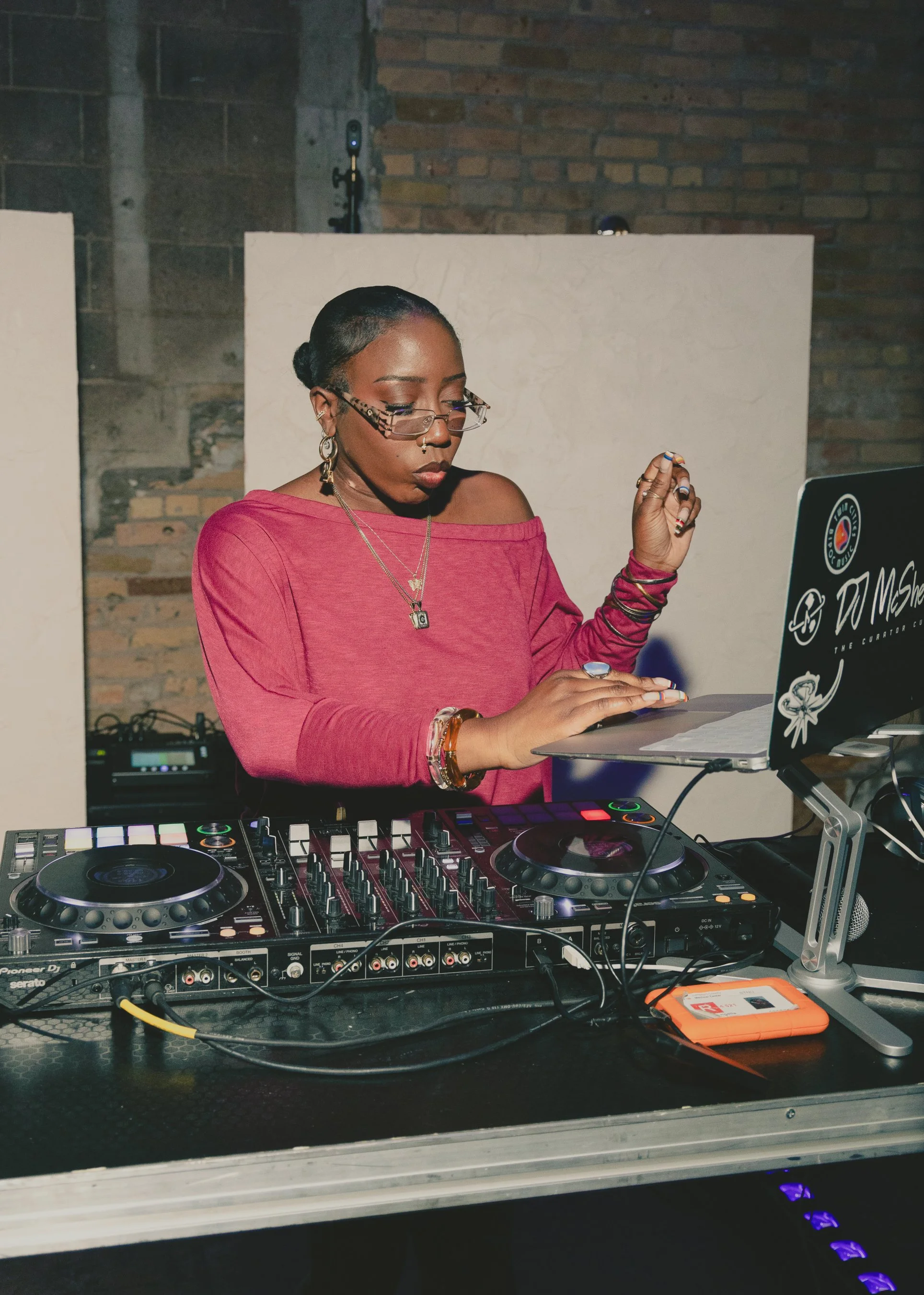 A female DJ is standing behind a DJ controller, wearing glasses and a pink off-the-shoulder top. She is focused on her equipment, with a laptop to her right displaying various stickers. The background includes brick walls and professional audio equip