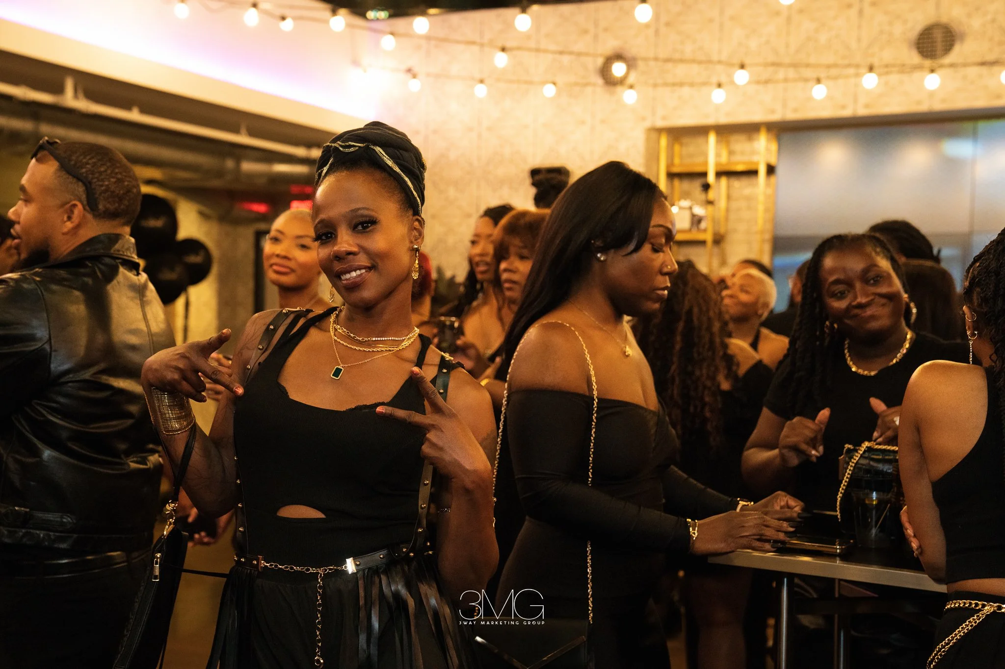 A group of women and men gathered in a dimly lit indoor space, likely at a social event or party. The women are dressed in black outfits and gold jewelry, with some smiling and posing for the photo. String lights are hanging above, creating a warm at