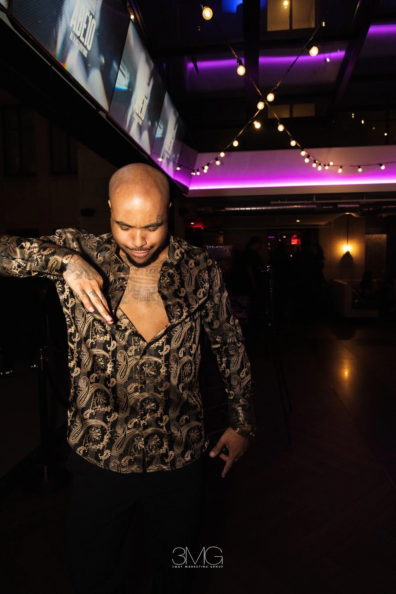 A man with tattoos on his face and chest, wearing a patterned shirt, is looking down and touching his neck. The background features purple lighting and a dimly lit room with hanging string lights. The logo '3MG' is visible at the bottom of the image.
