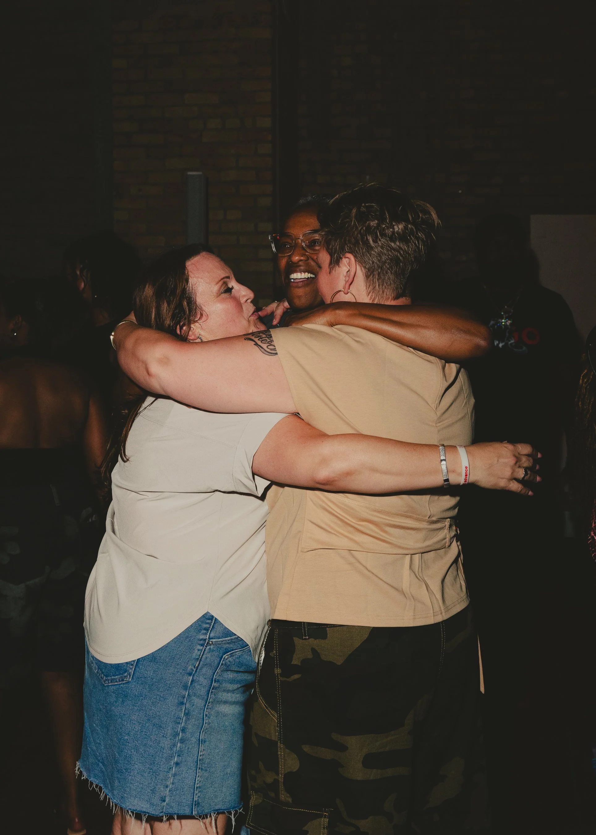 Two women hugging tightly and smiling at each other at a social event, with a third person smiling in the background, against a dark brick wall.
