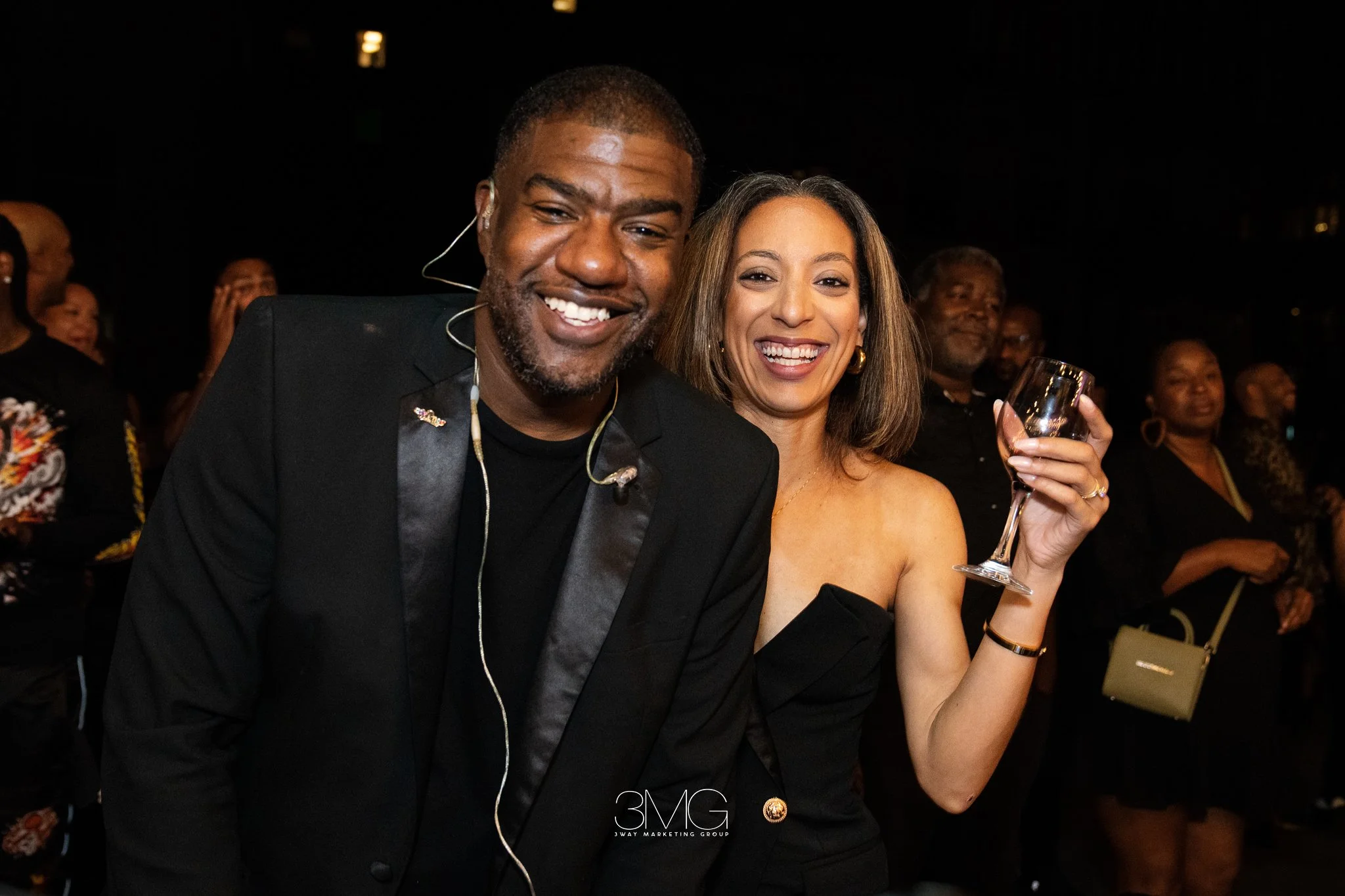 A smiling man in a black suit with a woman holding a wine glass at an indoor event. Both appear happy, surrounded by other attendees.
