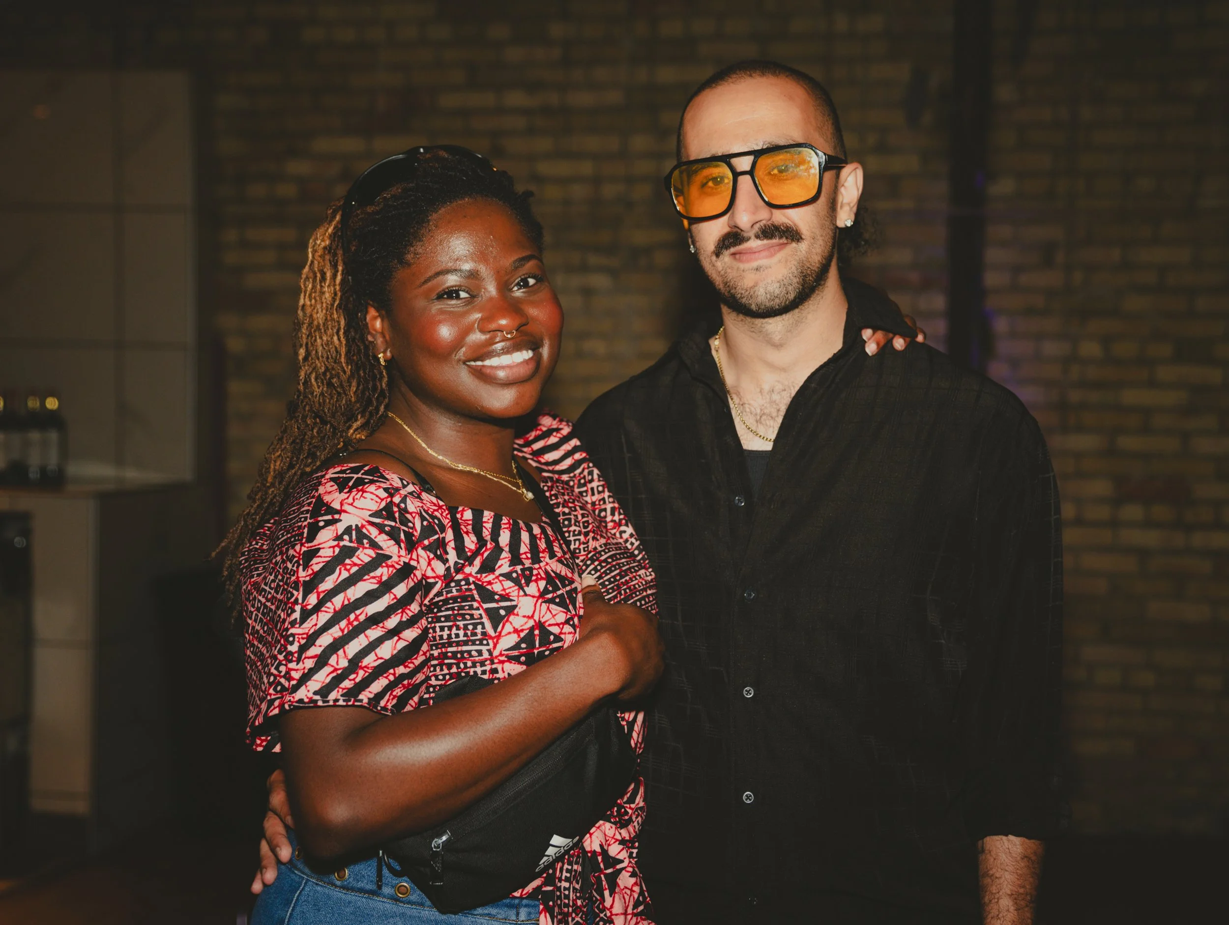 A woman and a man standing close together indoors, smiling at the camera. The woman has dark skin, dreadlocks, and is wearing sunglasses on her head, a white and red patterned top, and gold jewelry. The man has light skin, a beard, short hair, and is