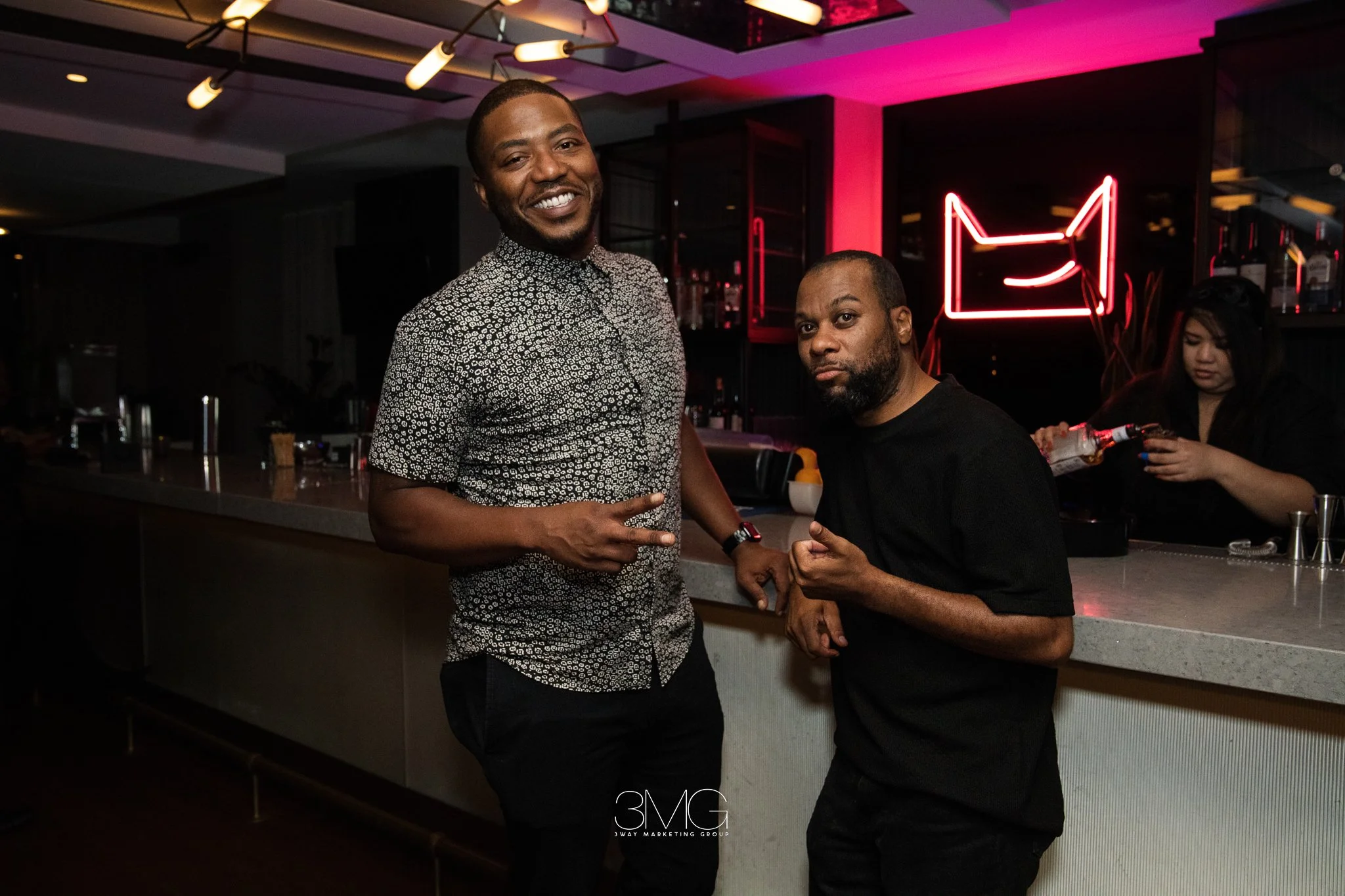 Two men posing for a photo at a bar, one smiling and making a peace sign, the other with a serious expression. There is a woman in the background preparing a drink, and a neon sign of a cat in the background.