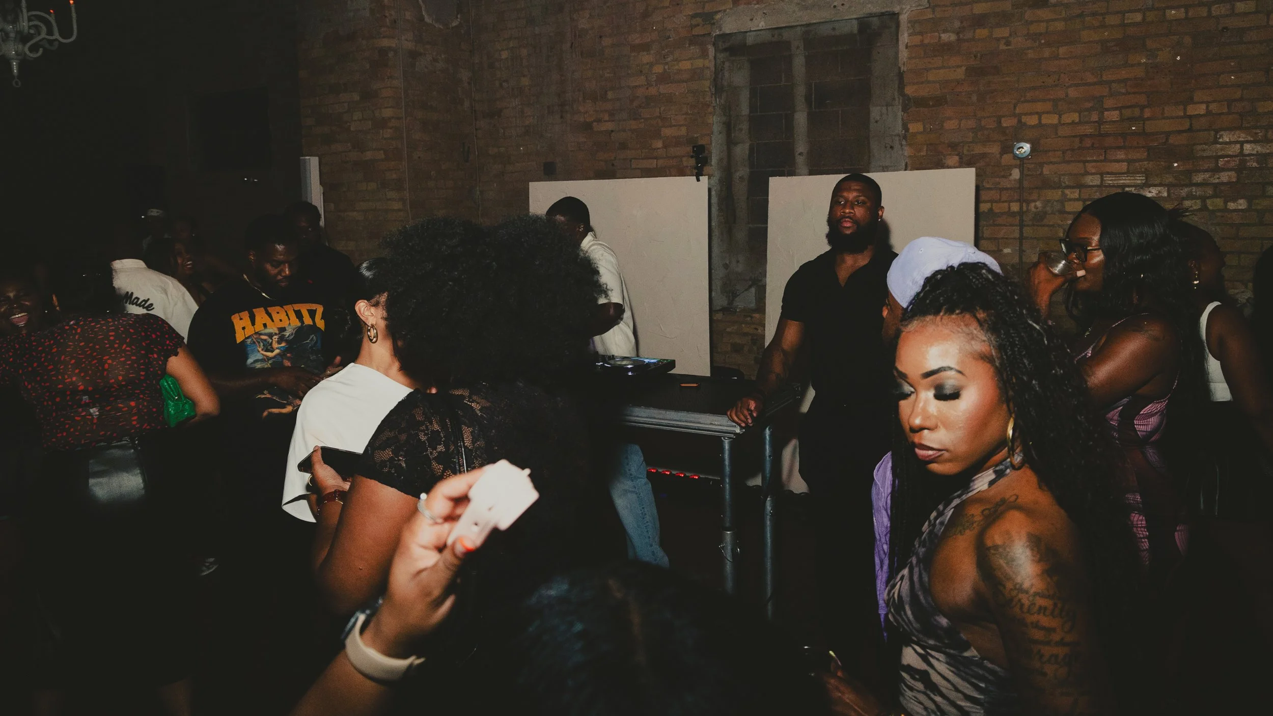 People gathered at a dimly lit indoor event, with a DJ playing music on turntables in front of a brick wall, some women with tattoos and curly hair, and others casually dressed, socializing and enjoying the atmosphere.