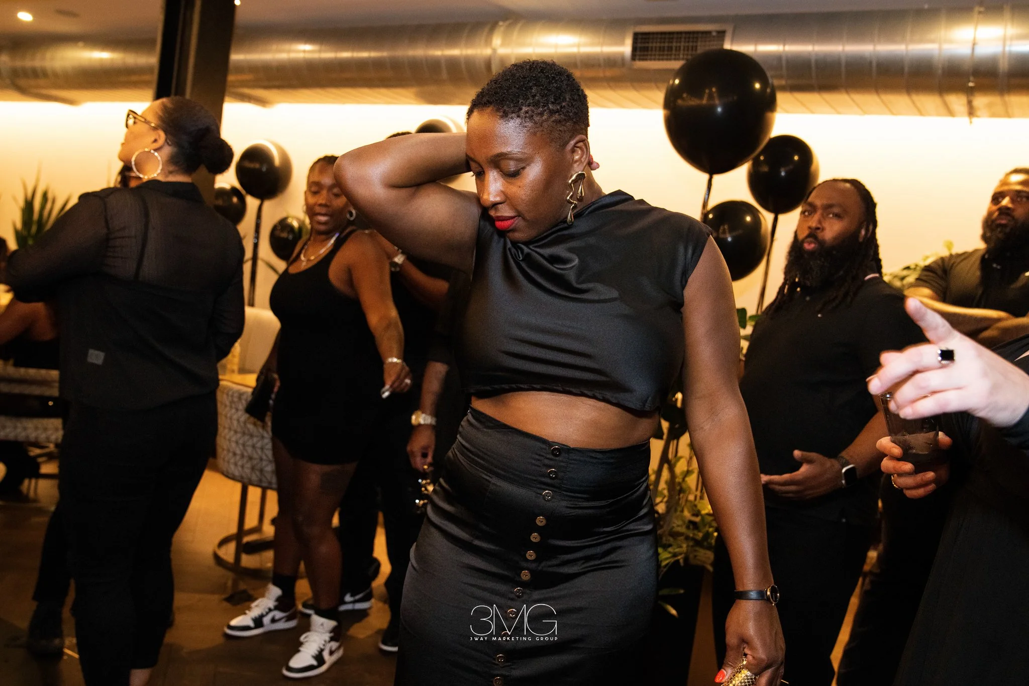 A woman with short curly hair wearing a black sleeveless top and high-waisted skirt, with red lipstick, is dancing and looking down at an event with other people. Behind her, other attendees are standing, some with balloons in the background, in a di
