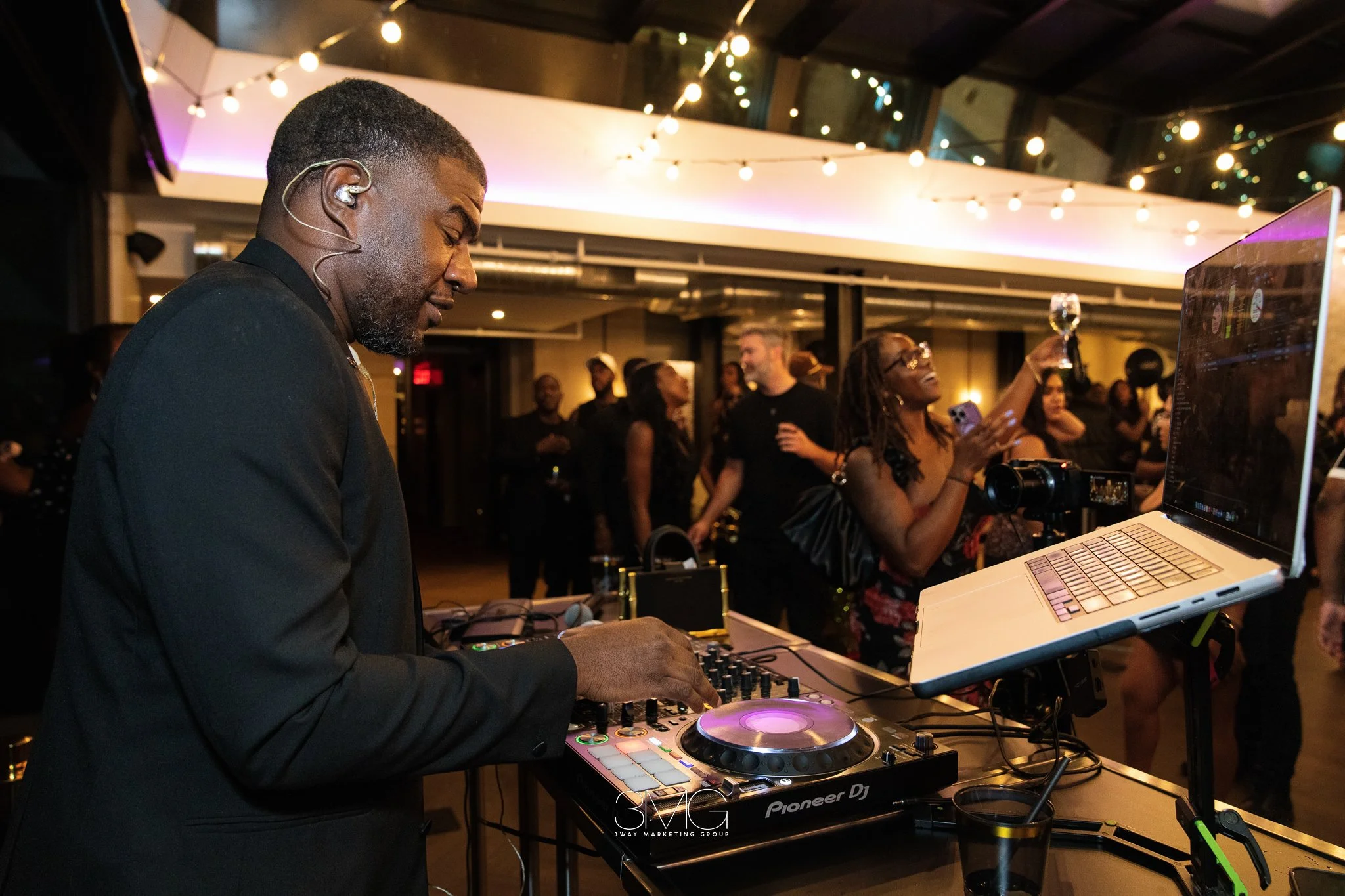 A man in a black jacket DJing at an indoor event with string lights and people dancing in the background.
