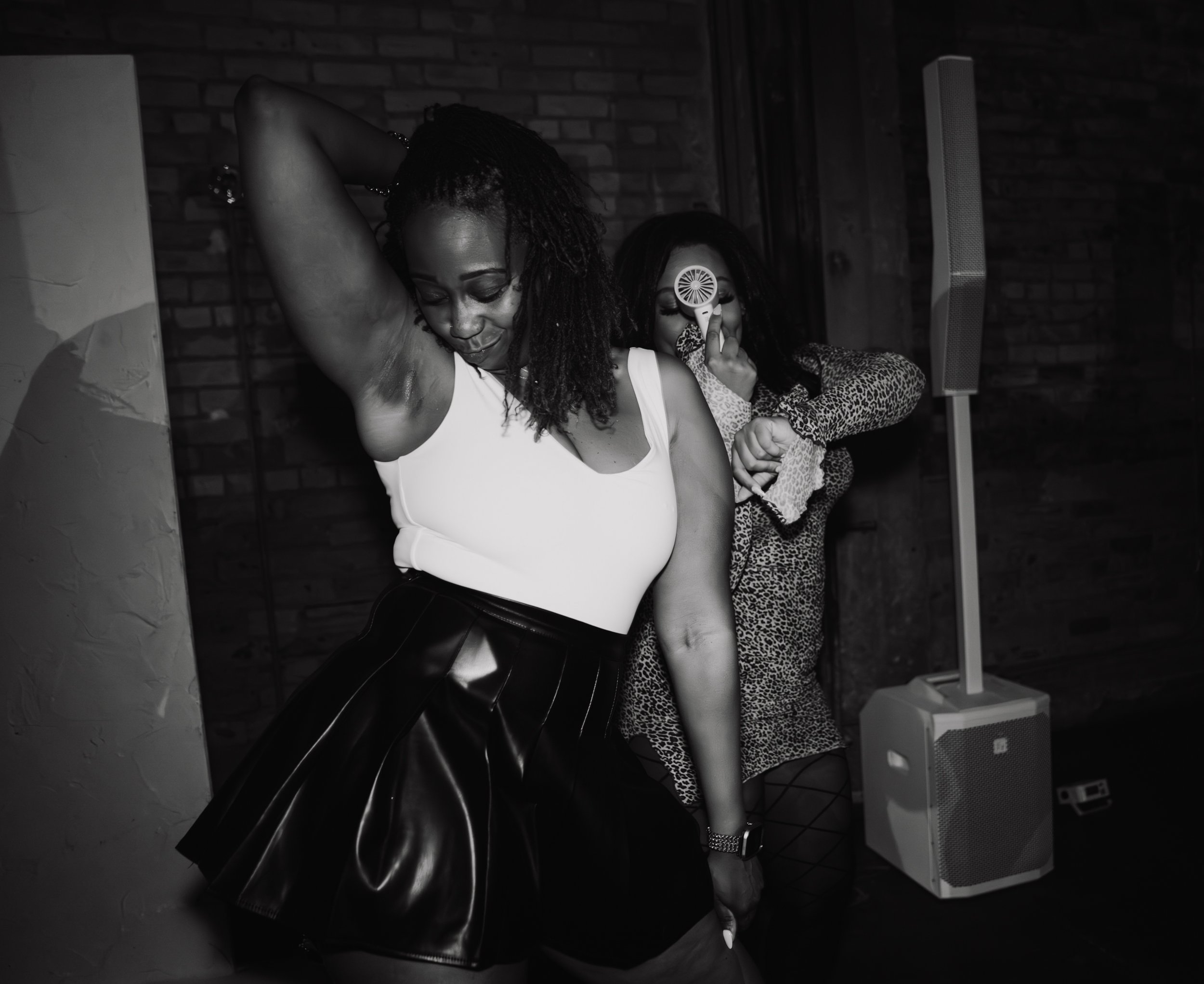 A woman with dreadlocks and a nose piercing dancing with her eyes closed in a dimly lit room, wearing a sleeveless top. Behind her, a person with a leopard print jacket is holding a handheld fan near their face. There is a speaker on the floor and a 