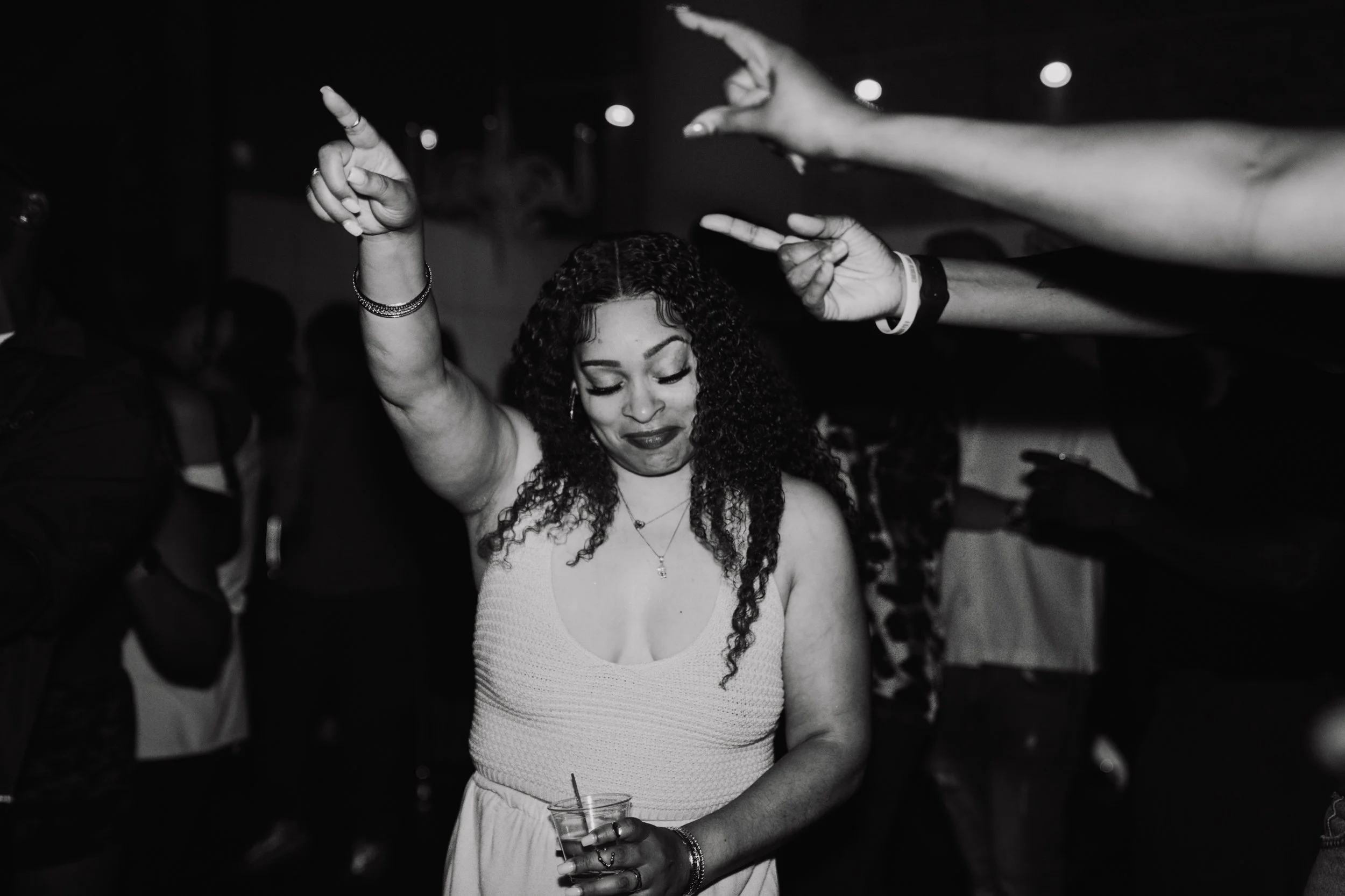 A woman with curly hair holding a drink with a straw and smiling with her eyes closed, surrounded by people at a party or dance event in a dimly lit environment.