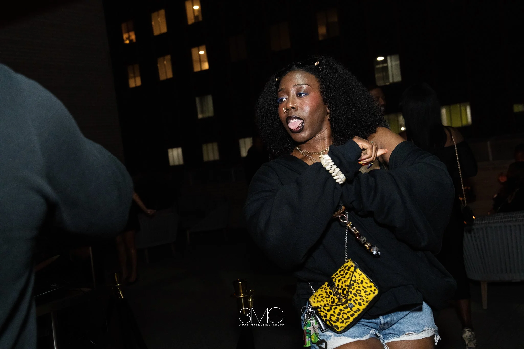 A woman with dark curly hair, makeup, and jewelry, sticking her tongue out, at night-time outdoor event, with buildings and other people in the background.