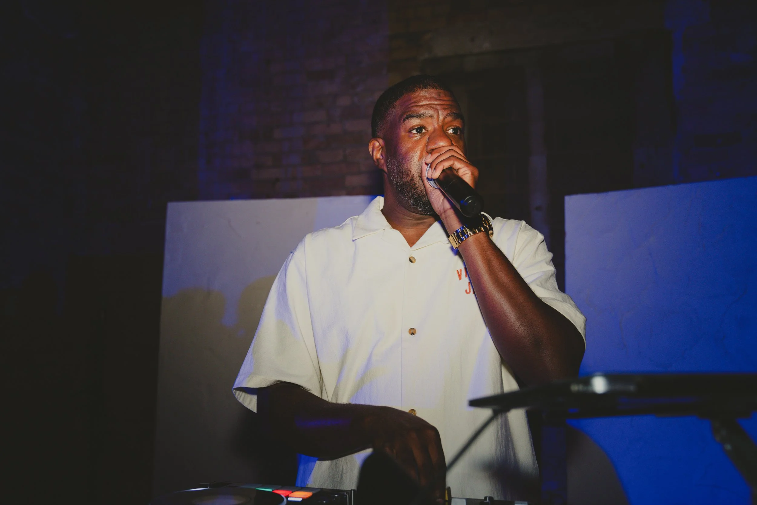 A man with a beard and short hair, wearing a white shirt and a gold wristwatch, holding a microphone and performing on stage with a dark brick wall background.