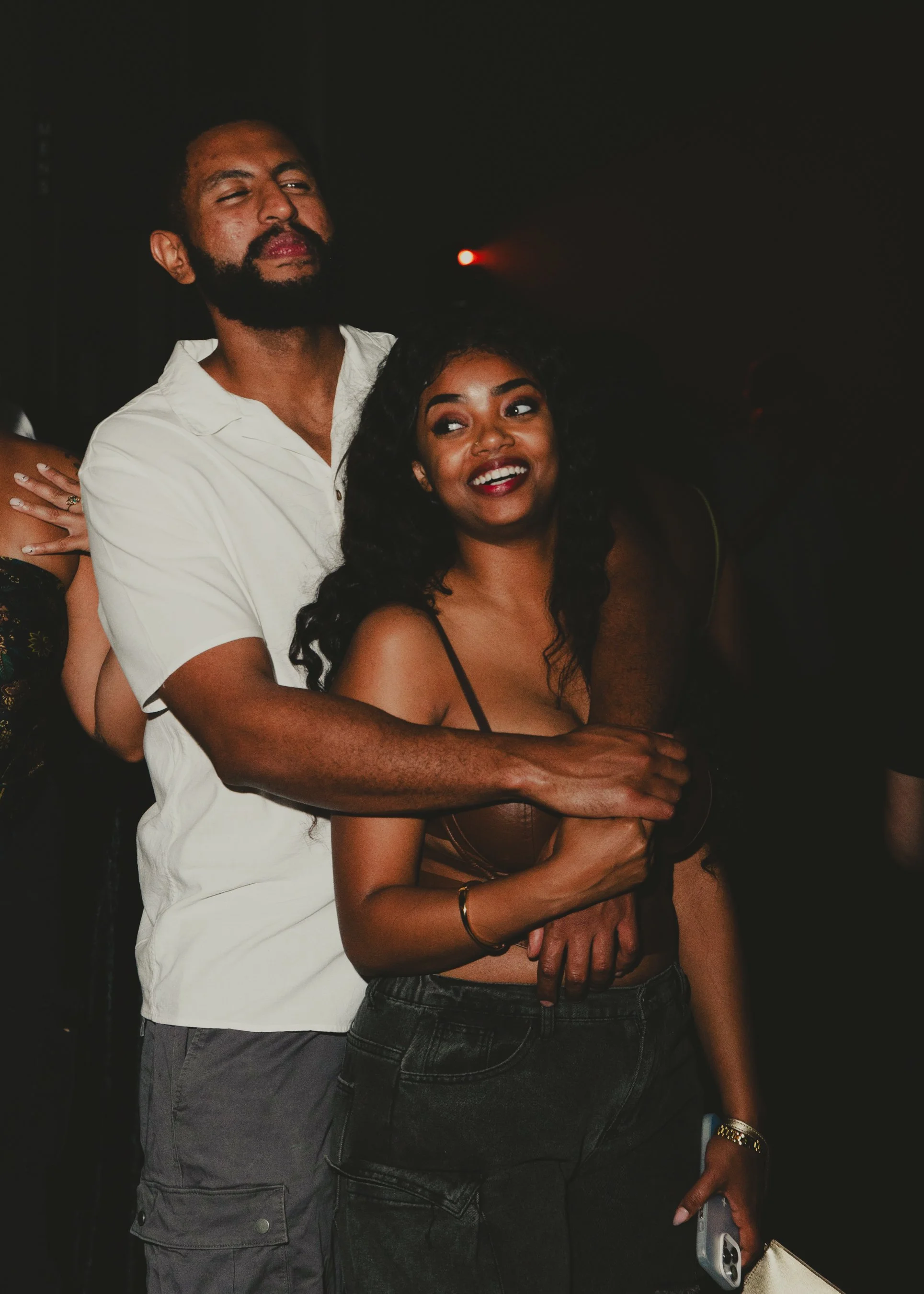 A man and woman standing closely together at a dimly lit event, with the man holding the woman from behind as they look off to the side, smiling.